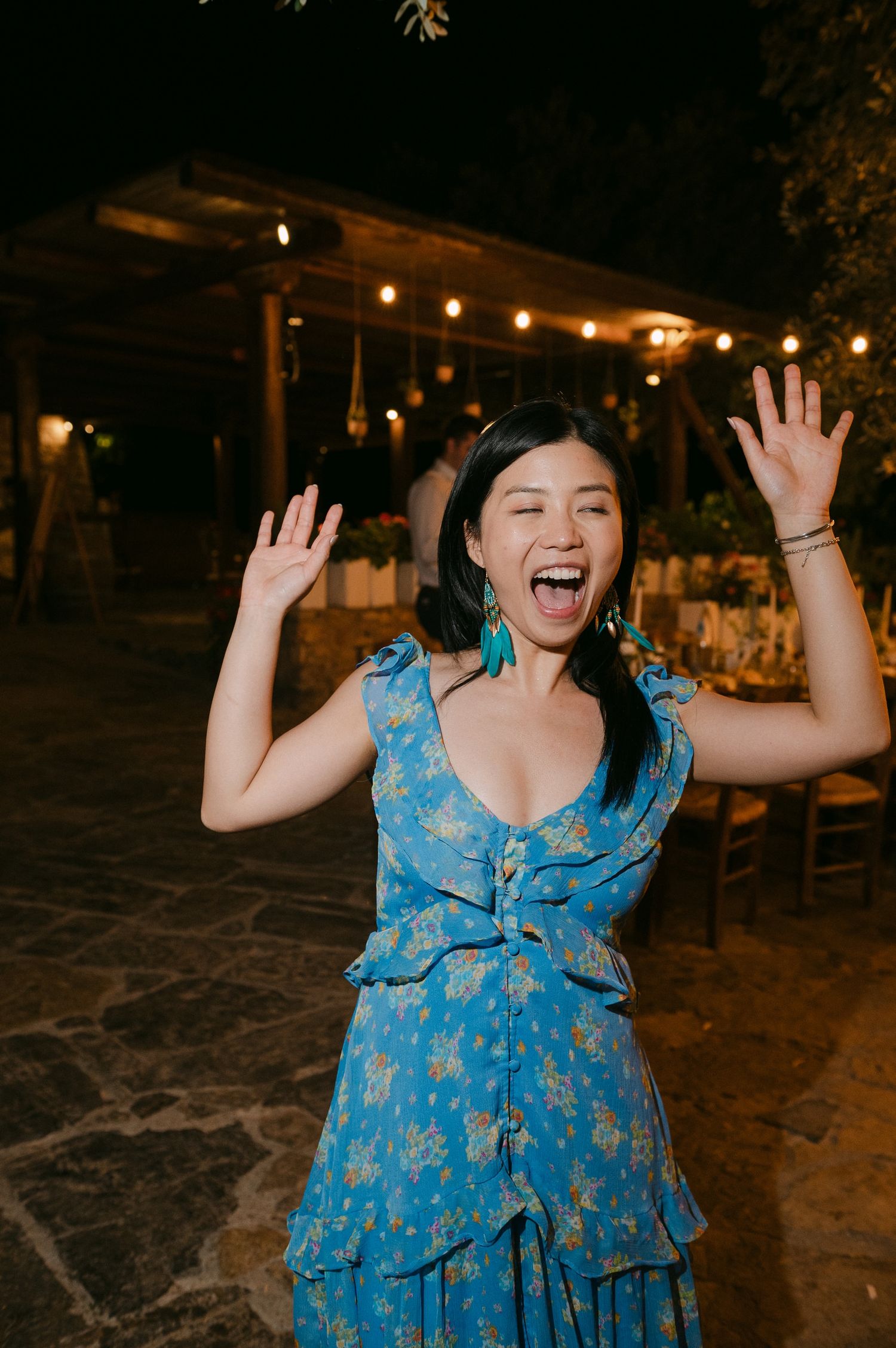 A joyful figure in a blue floral dress with ruffled tiers dances and twirls at an outdoor evening event.