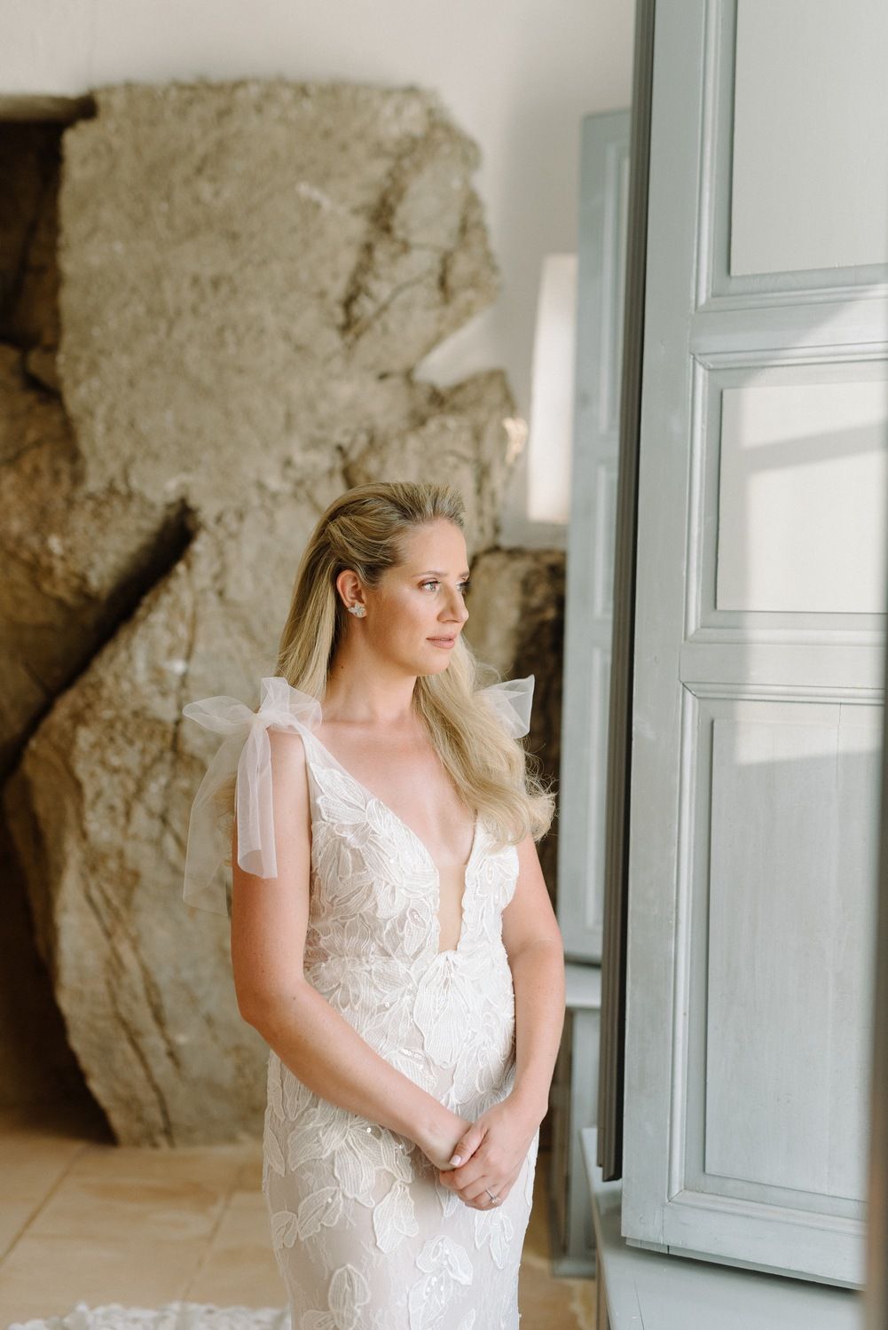 A person in a detailed lace wedding dress with sheer sleeves stands in elegant pose by a window.
