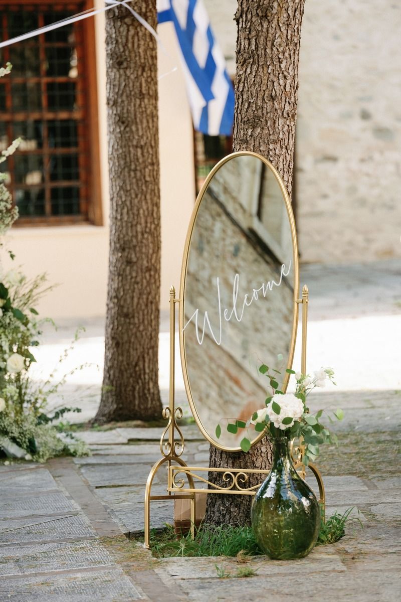Elegant gold-framed welcome mirror display with floral arrangements at an outdoor wedding venue.