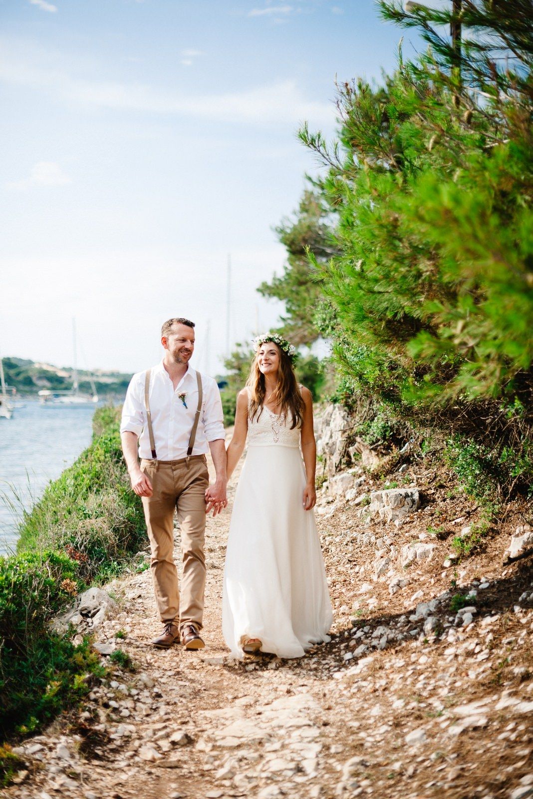 Couple in bohemian wedding attire walking along coastal path with ocean views.