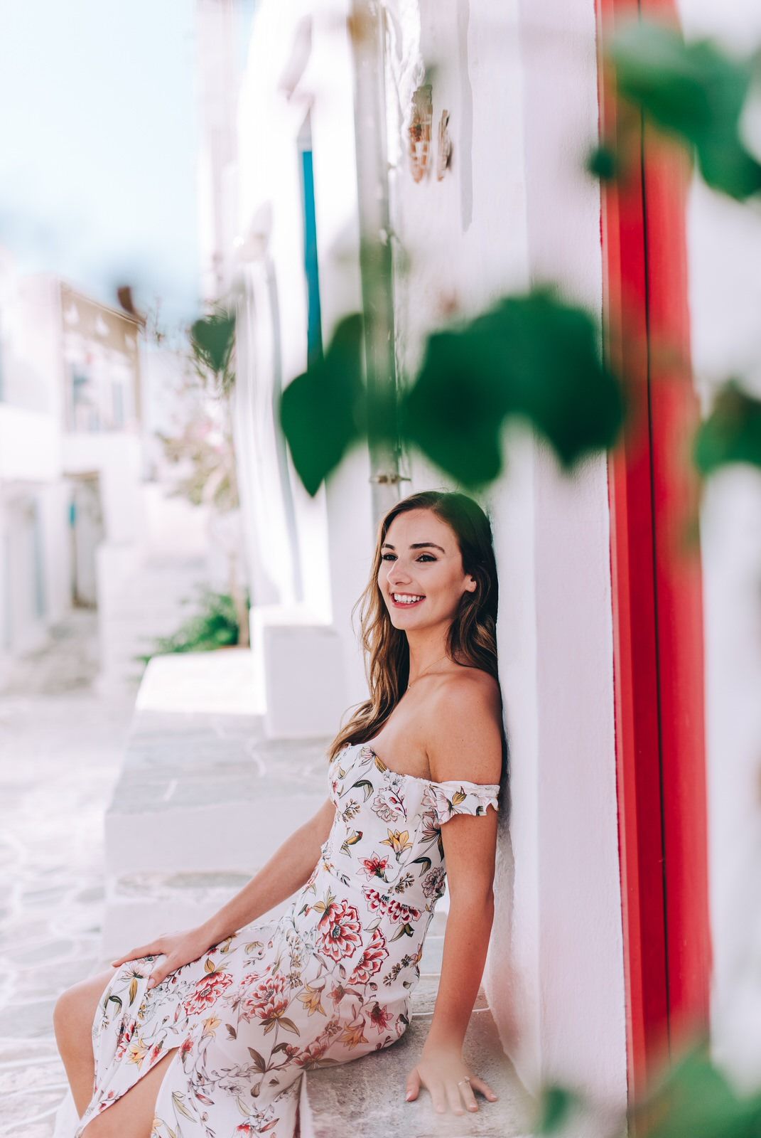 Woman in off-shoulder floral dress poses against white wall with red accents.