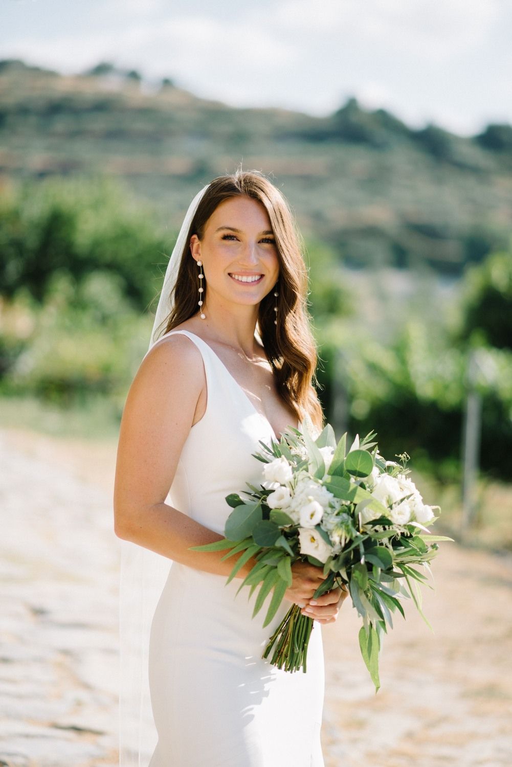Bride in elegant white dress holding a green and white bouquet against a hillside landscape.