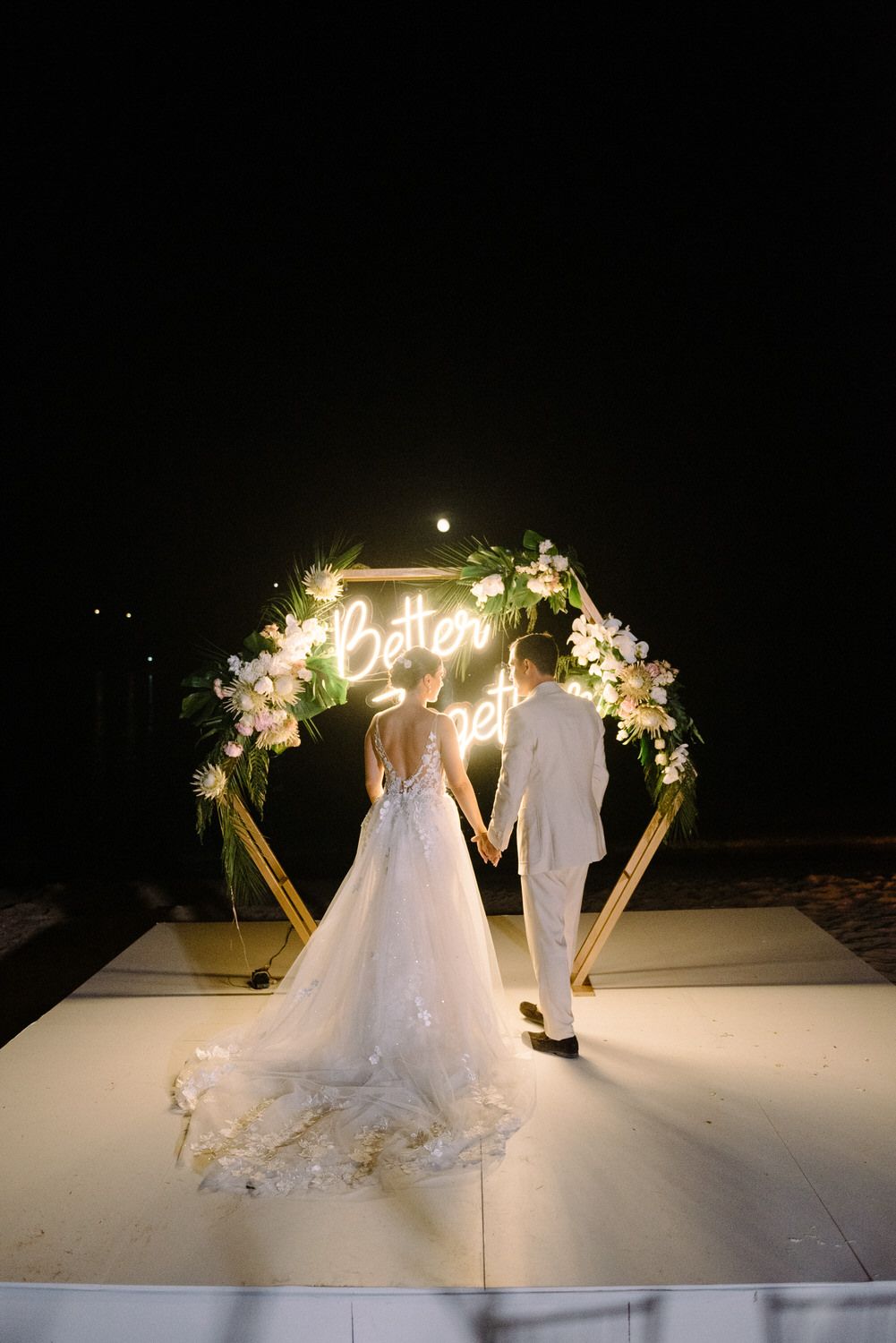 A wedding couple stands beneath an illuminated 'Better Together' neon sign arch decorated with white flowers at night.