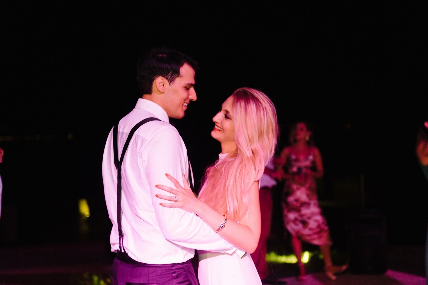 A romantic couple dancing together at an evening celebration with pink lighting.