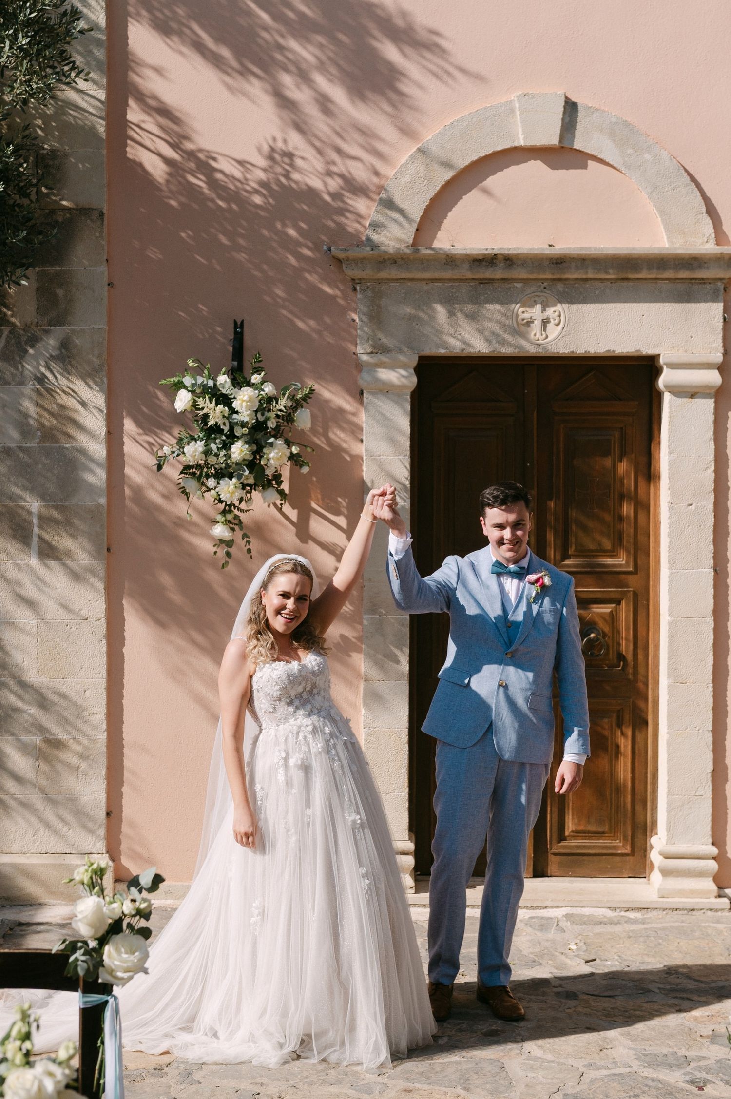 A wedding couple poses joyfully outside a sun-dappled Mediterranean church with white walls and wooden doors.