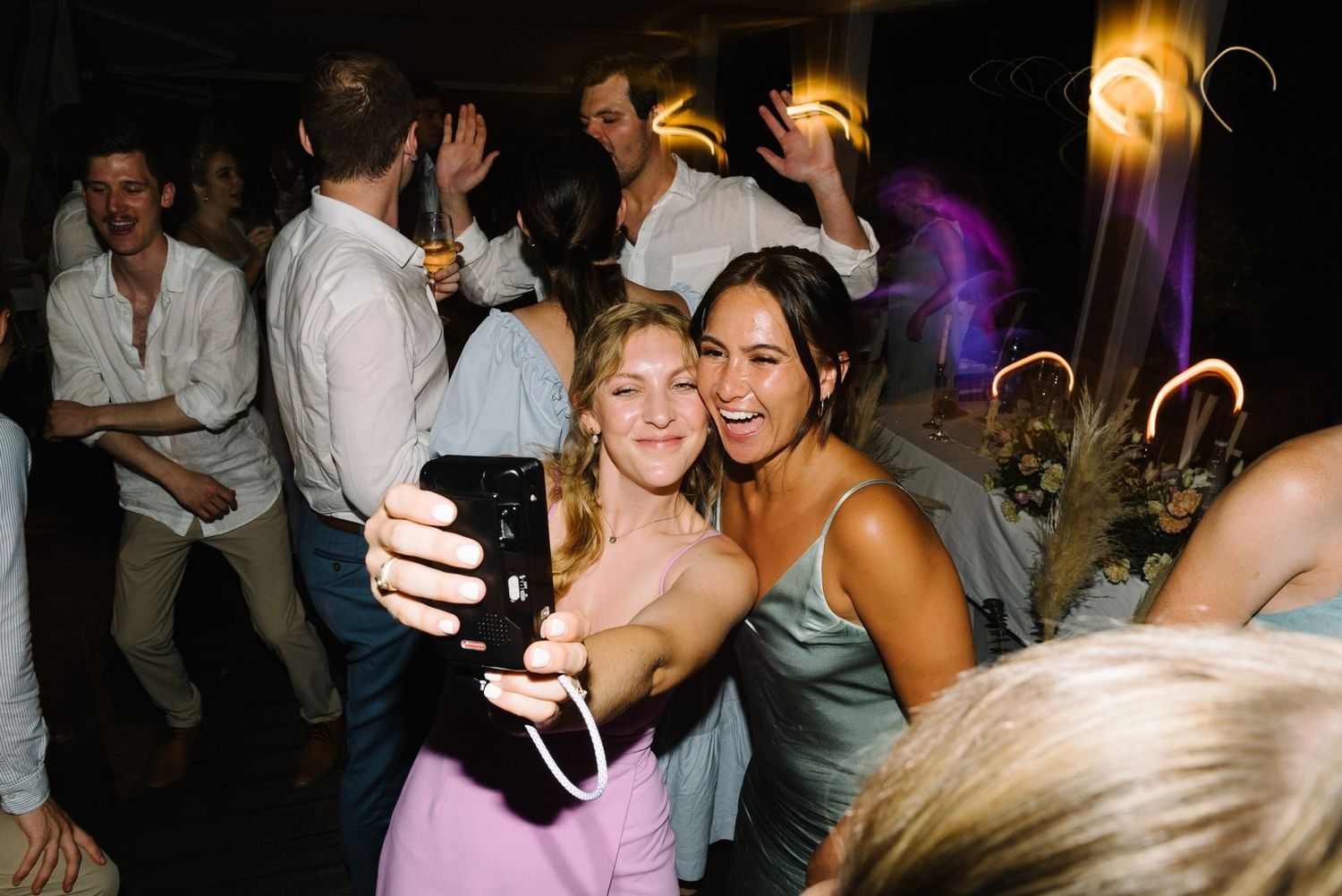 Two party guests take a selfie together while dancing at a nighttime celebration.