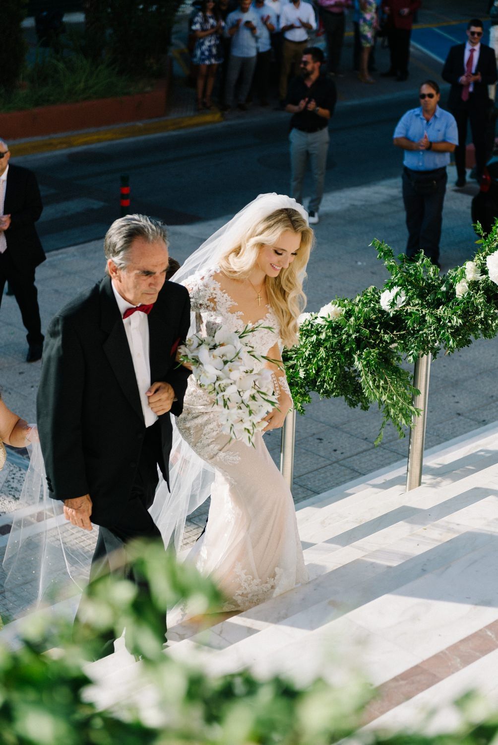 Elegant wedding procession walking down steps outdoors with flowing dress and floral decorations.