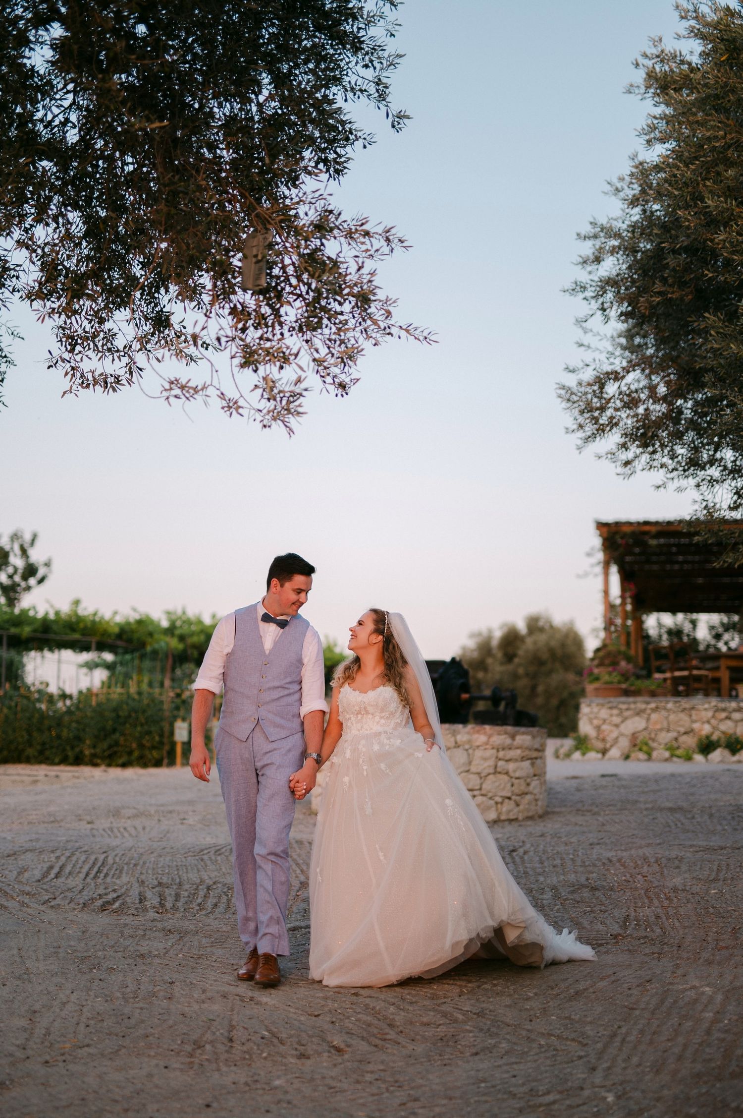 A romantic outdoor wedding scene at sunset with a couple walking together on a stone path beneath overhanging trees.