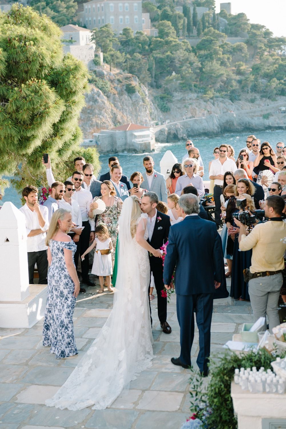 A beautiful outdoor wedding ceremony taking place on a stone terrace overlooking a Mediterranean coastal hillside.