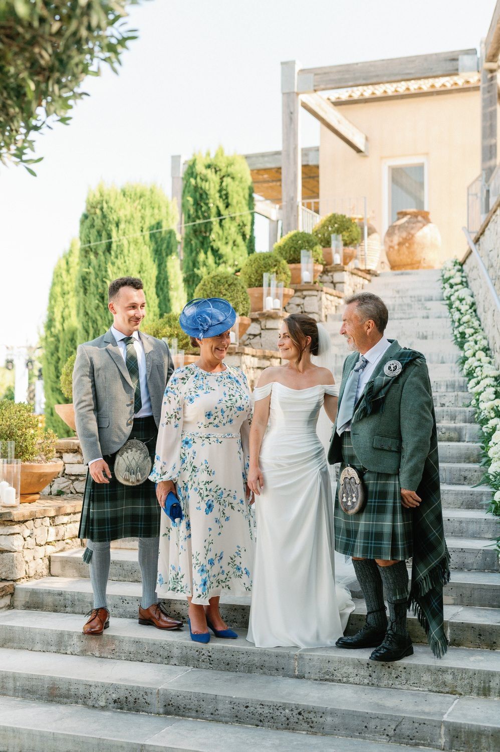 A wedding party in Scottish attire poses on stone steps with greenery and a peach-colored villa backdrop.