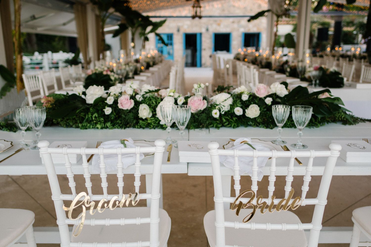 White reception chairs with 'Groom' and 'Bride' signs at an elegant wedding table setting with floral centerpieces.