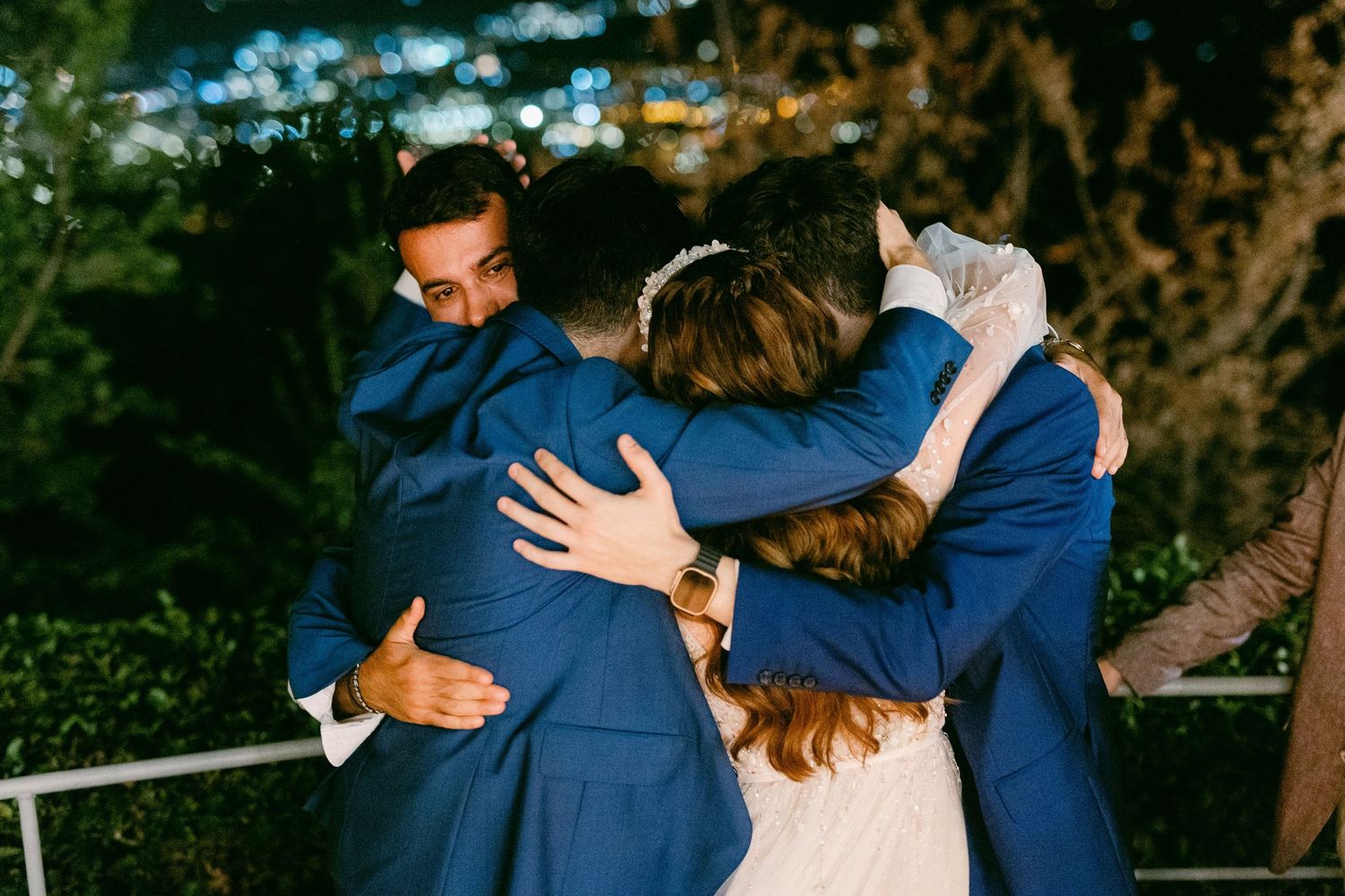 A group of people in navy blue suits share an emotional embrace outdoors during golden hour lighting.