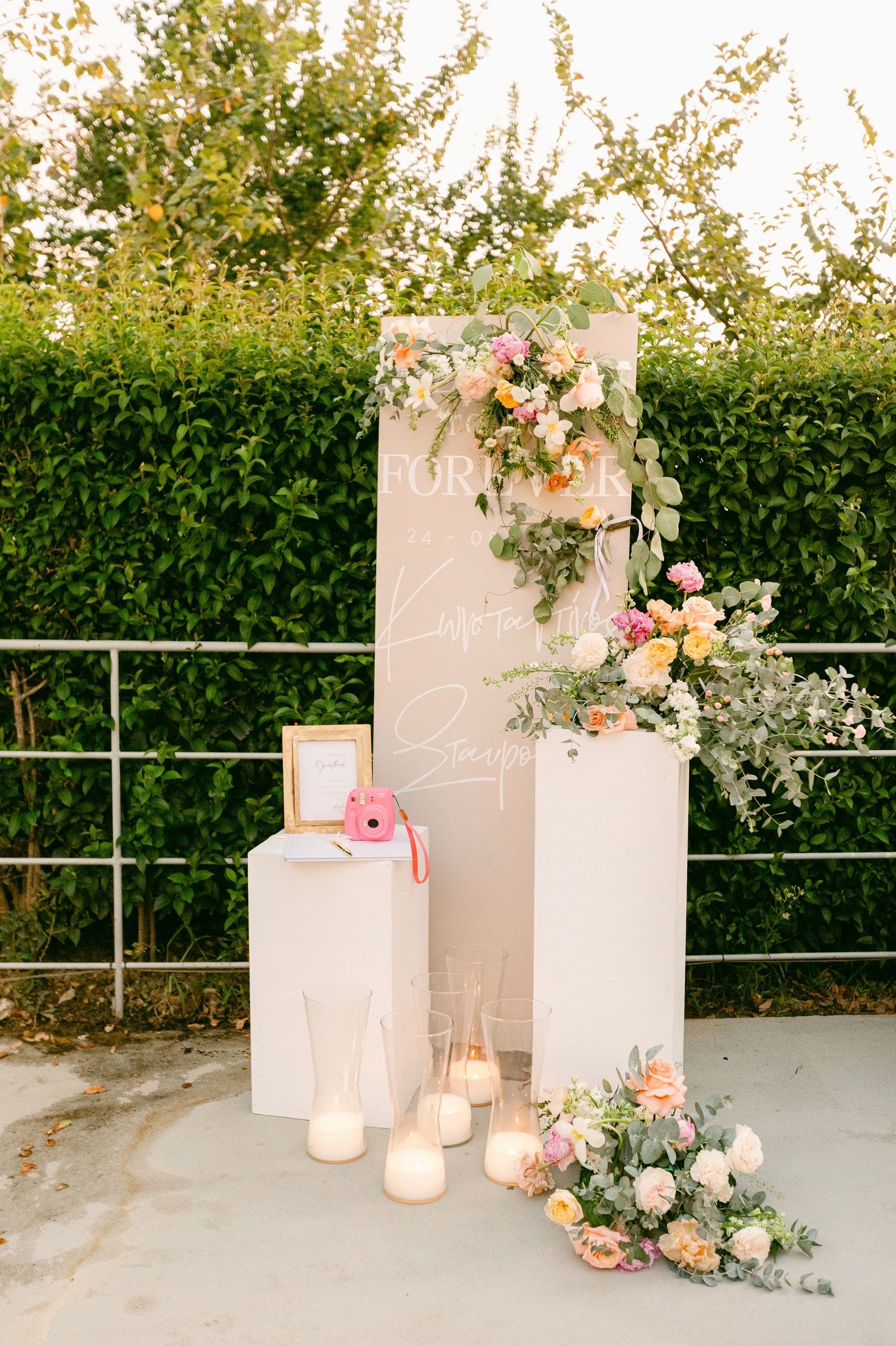 Elegant white pedestals decorated with peach and pink floral arrangements and candles against a garden hedge backdrop.