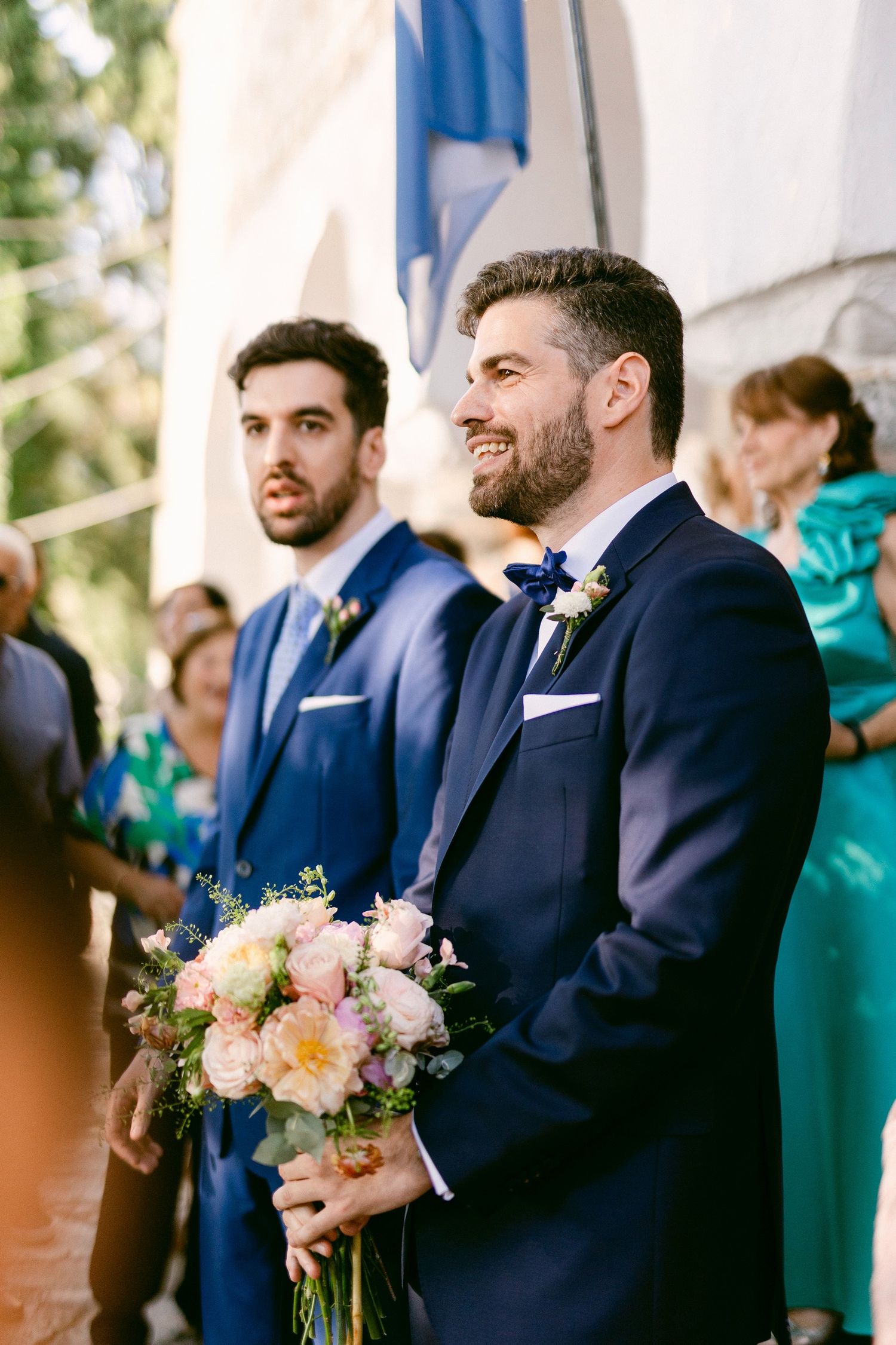 Two groomsmen in navy blue suits stand at a ceremony with one holding a pink and white floral bouquet.