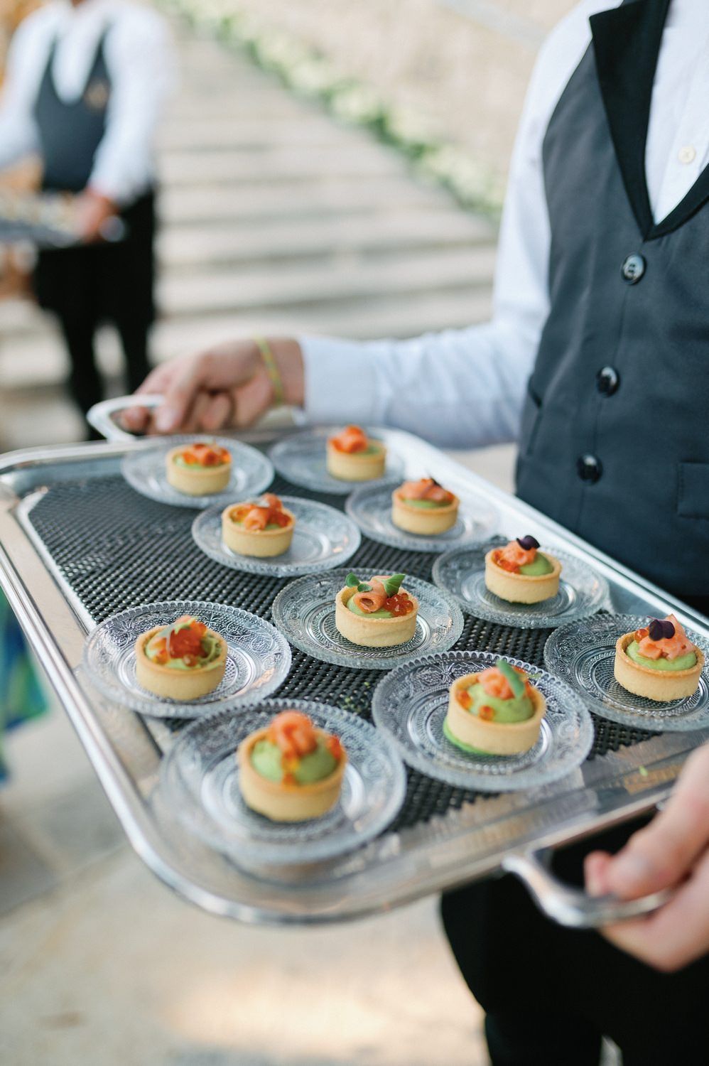 Server carries a tray of elegantly plated appetizers with garnishes on crystal plates at an upscale event.
