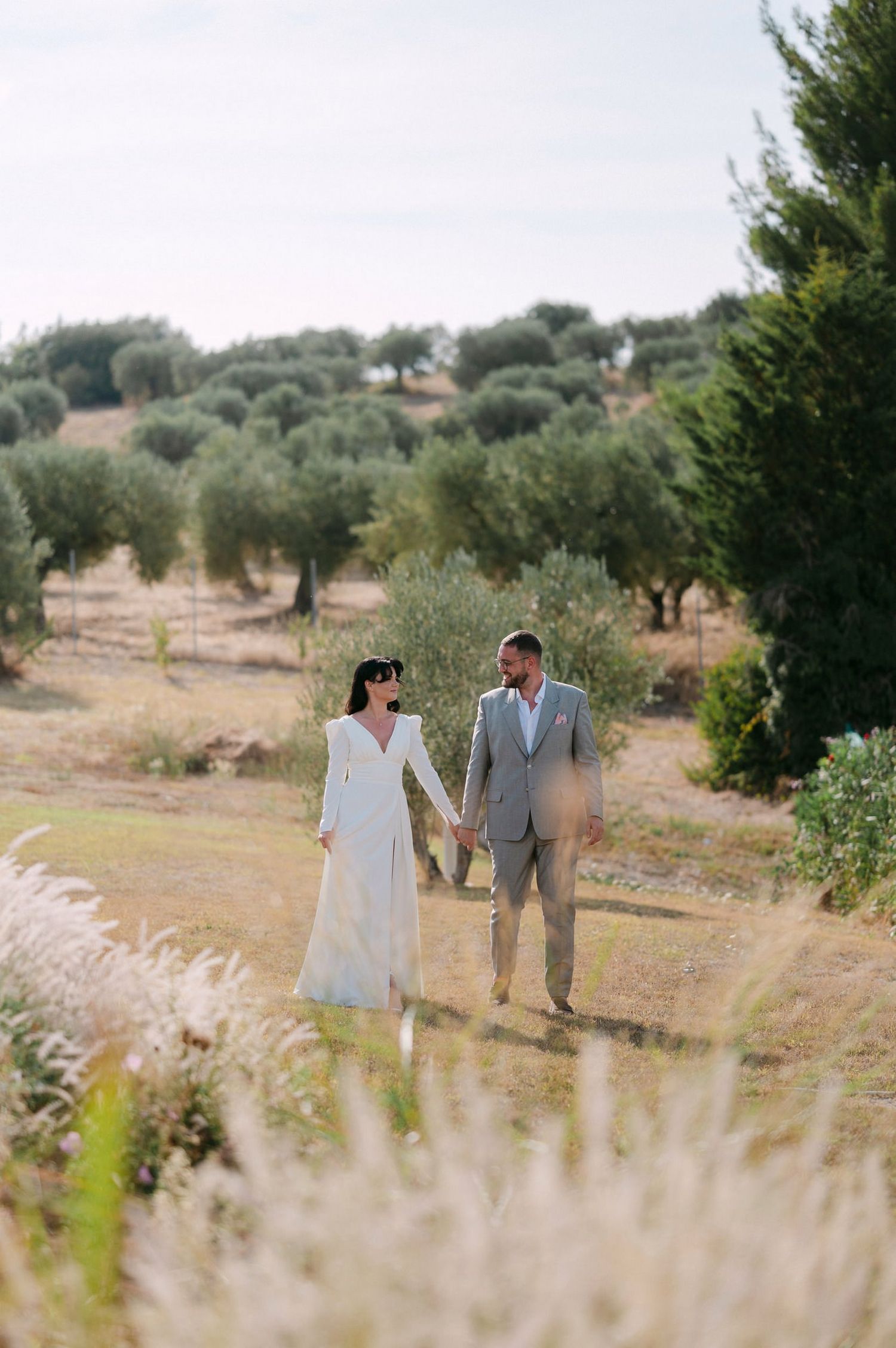 A couple walks hand in hand through a scenic olive grove on a sunny day in the countryside.