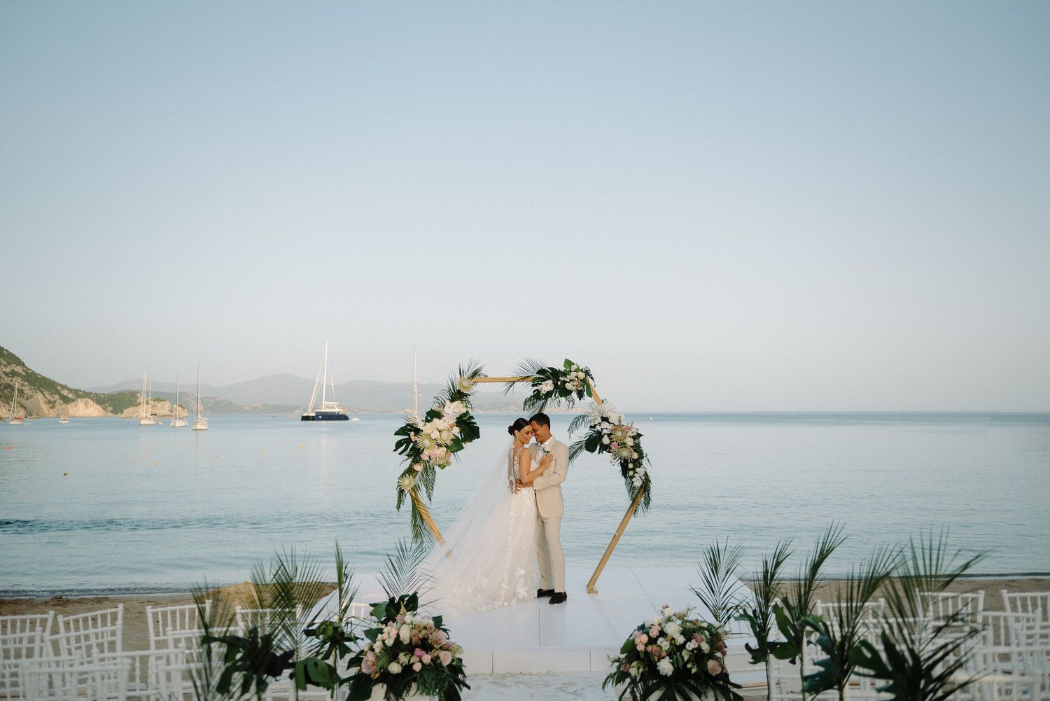 Wedding ceremony setup with floral arch and ocean backdrop at sunset on beach.