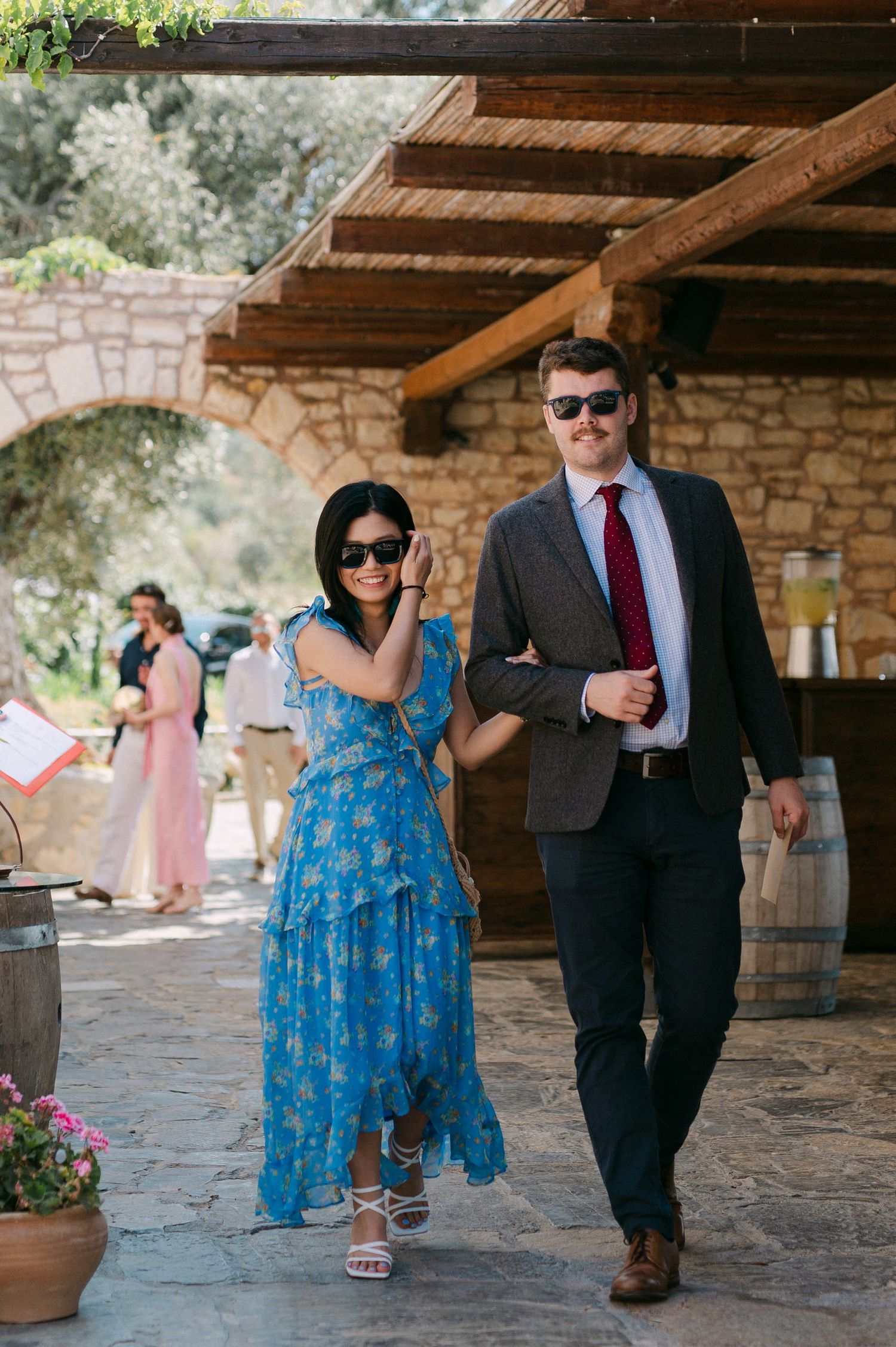 A couple in formal attire walks together under a covered stone patio with wooden beams.