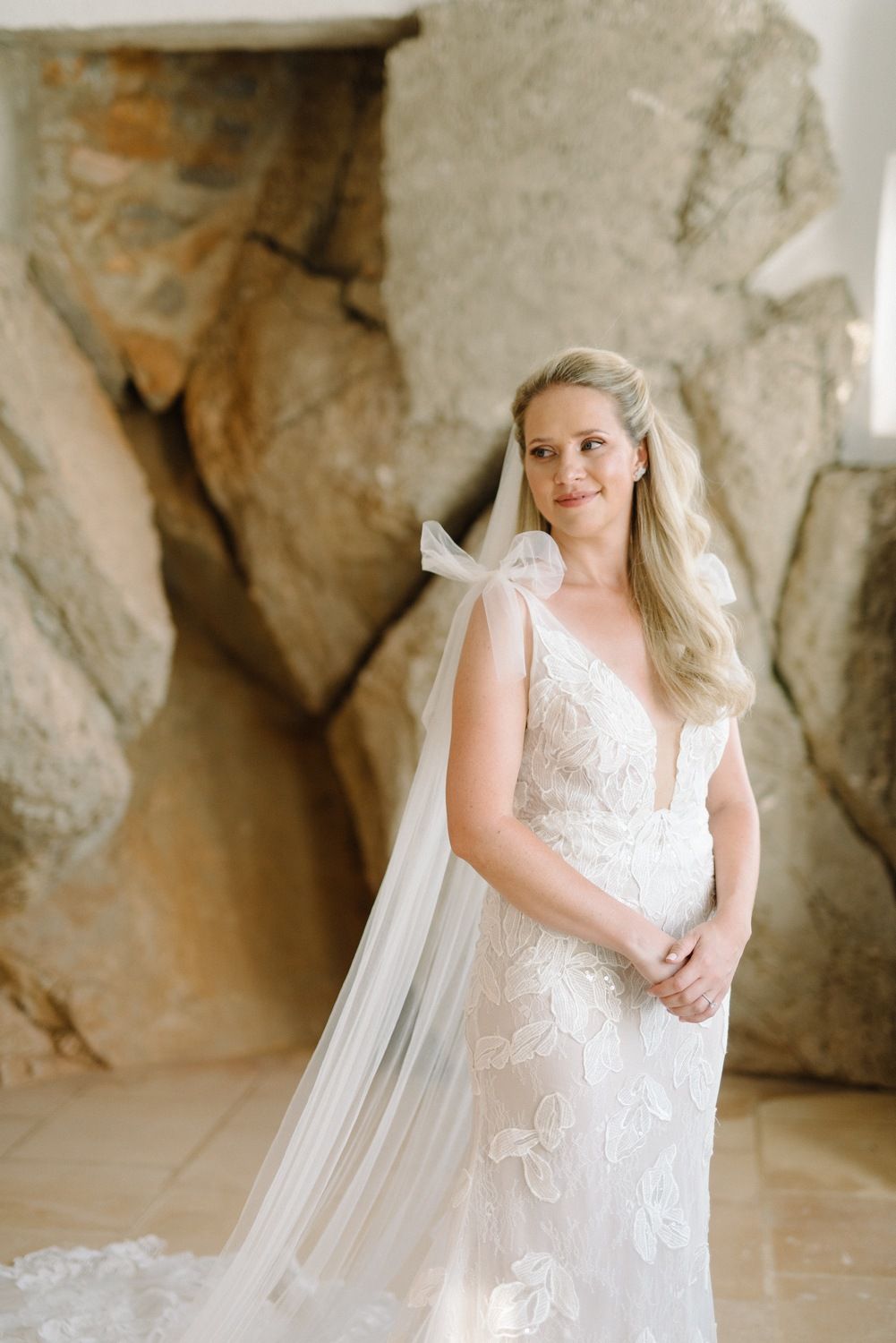 A person in a flowing lace wedding dress poses against a stone wall backdrop.