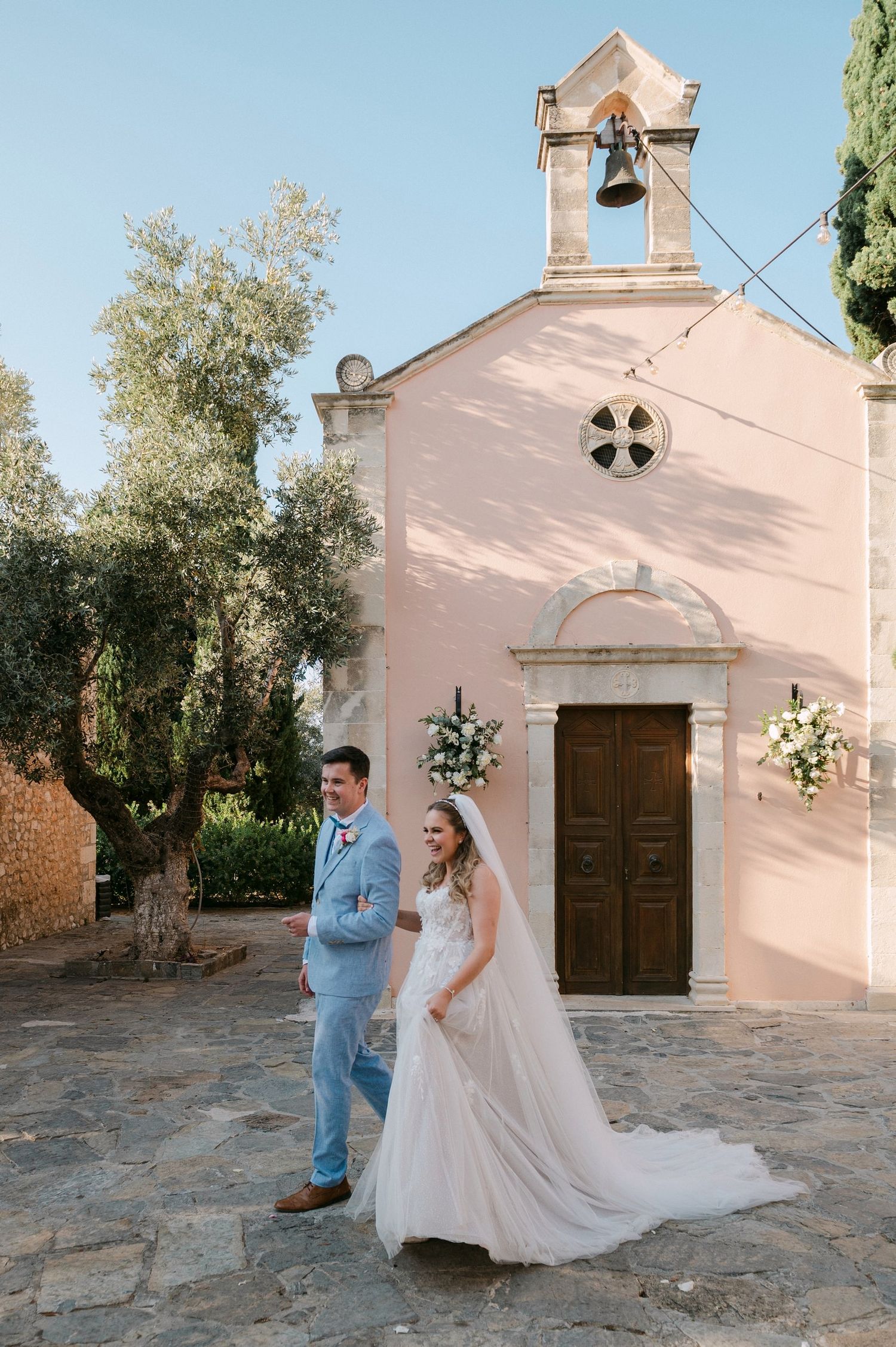 A couple celebrates their wedding day in front of a charming pink Mediterranean chapel with olive trees in the background.