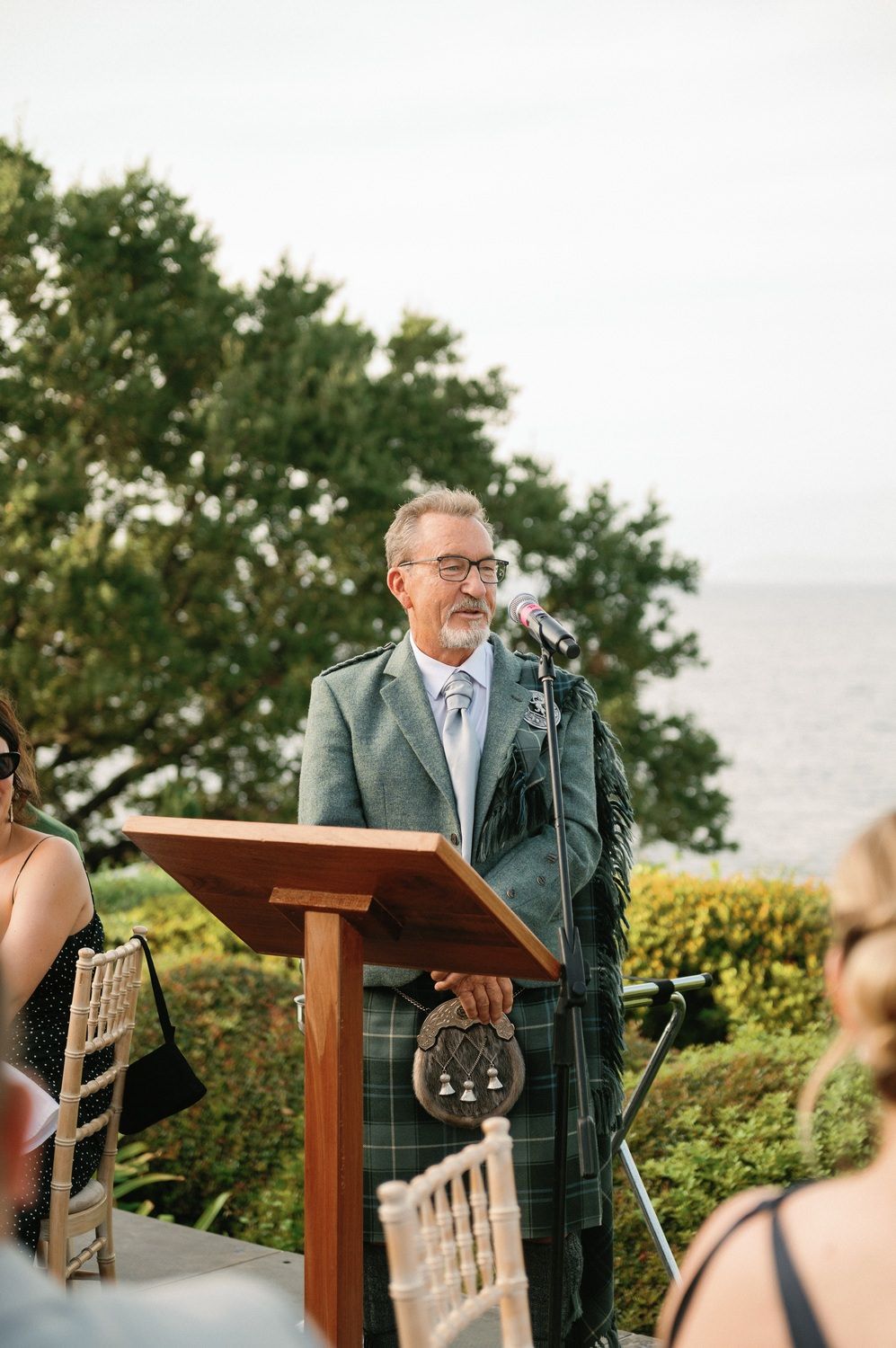 Speaker in traditional Scottish attire giving a speech at outdoor wedding ceremony.