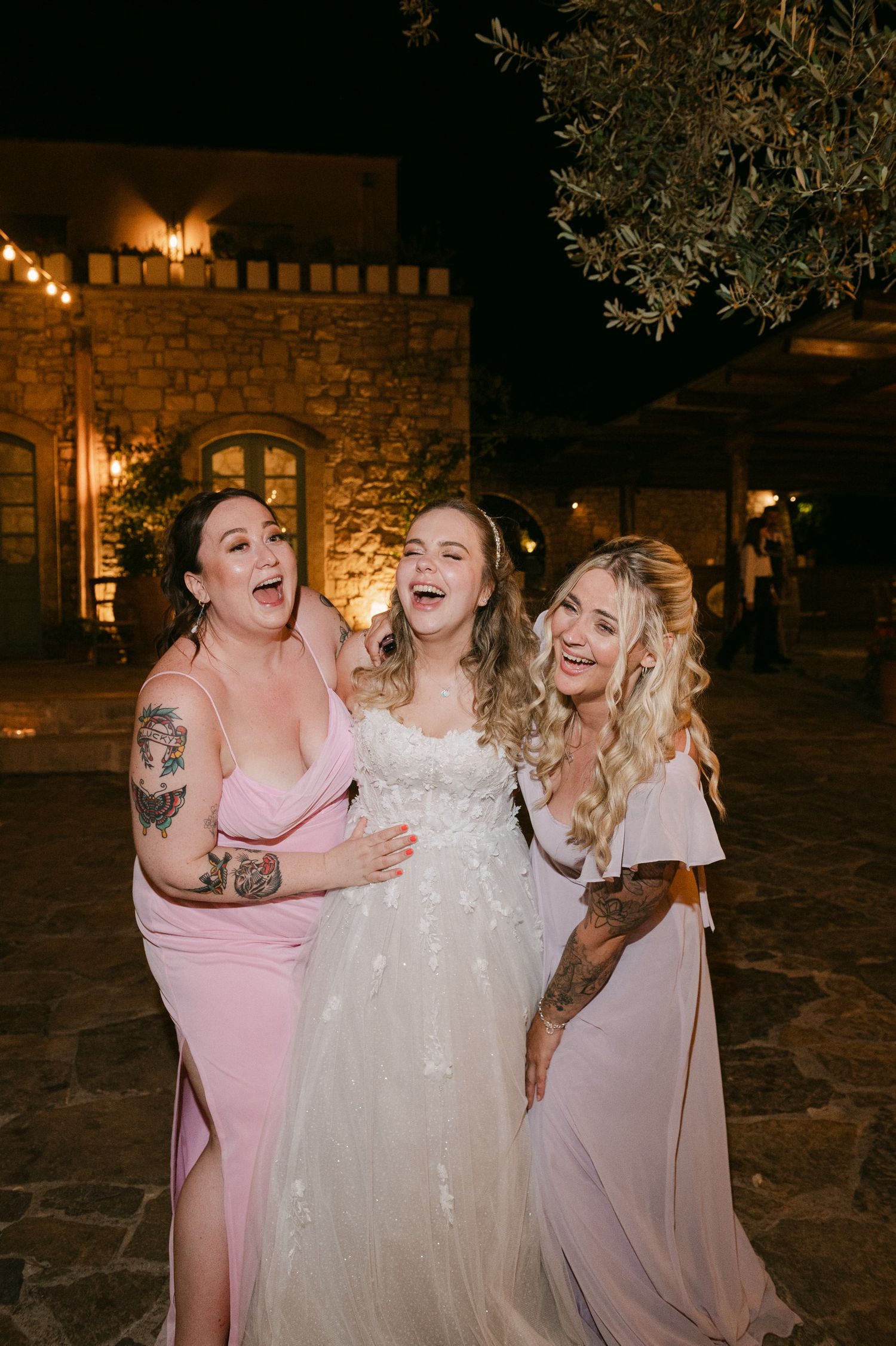 Three women in formal dresses share a joyful moment together at an outdoor evening wedding reception with stone walls and lights.