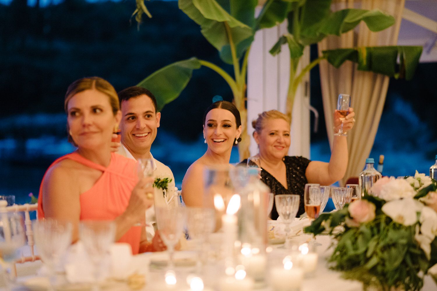 Evening wedding reception with guests seated at a candlelit table decorated with floral arrangements.