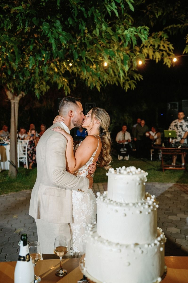 A couple shares their first kiss beside a three-tiered white wedding cake under garden lights.