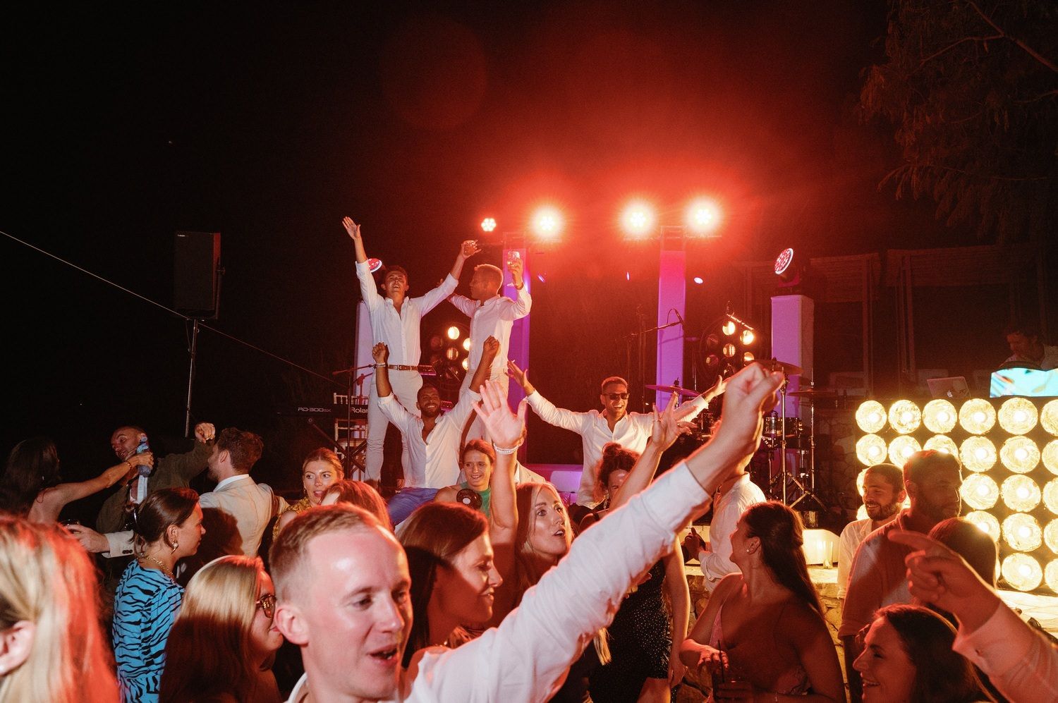 Dancing party-goers cheering with arms raised under red stage lights.
