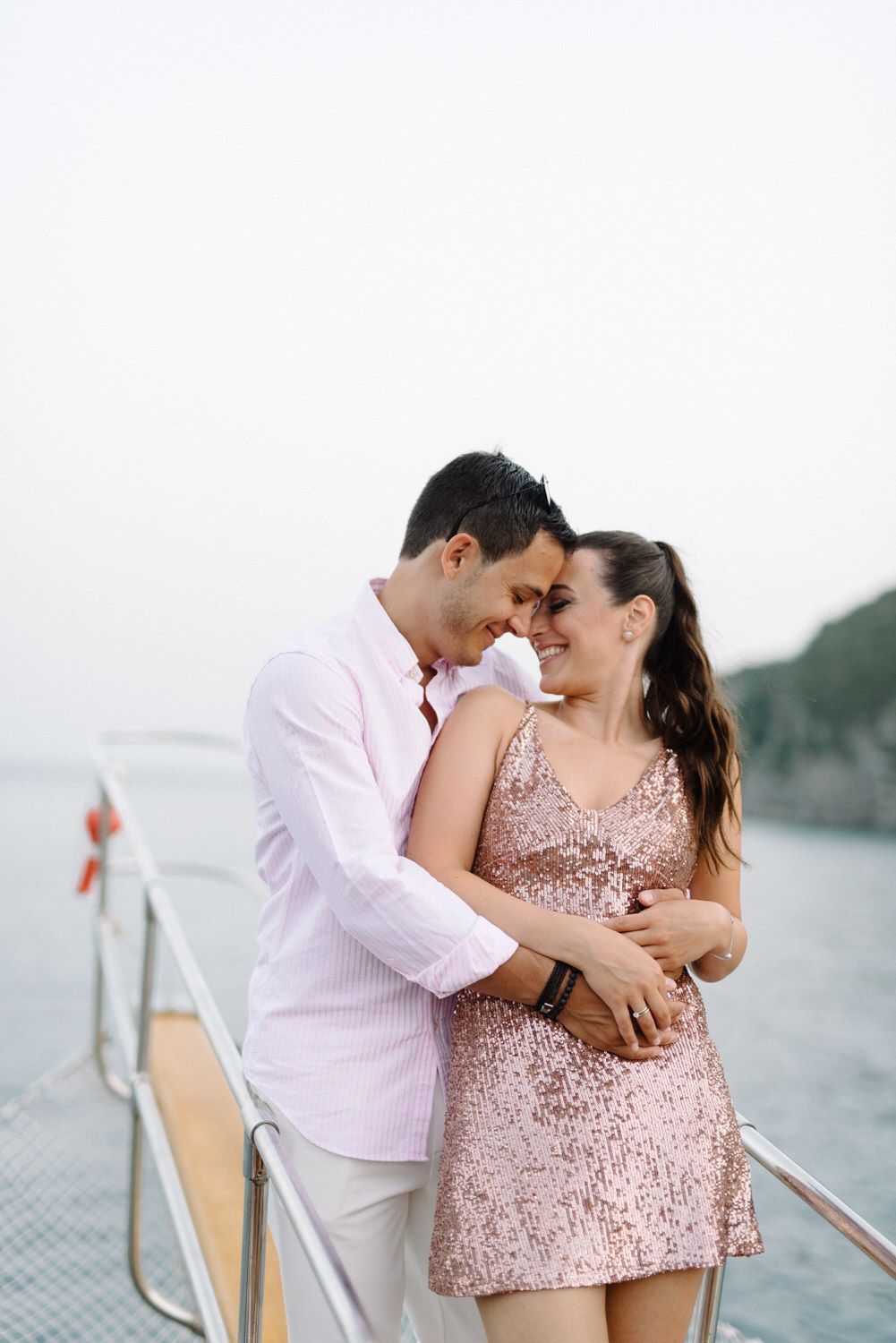 Couple embracing intimately on a yacht deck wearing coordinated pink and white outfits against a scenic backdrop.