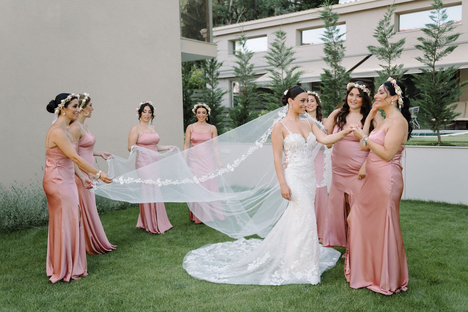 Bridesmaids in pink gowns arrange the bride's cathedral length veil in an outdoor garden setting.