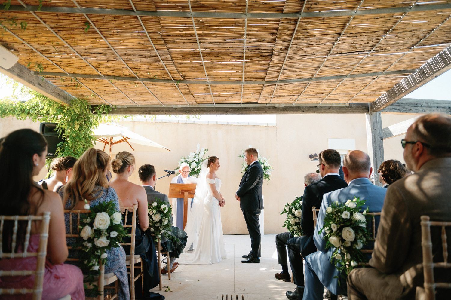 Outdoor wedding ceremony under a covered patio with guests seated watching the couple exchange vows.