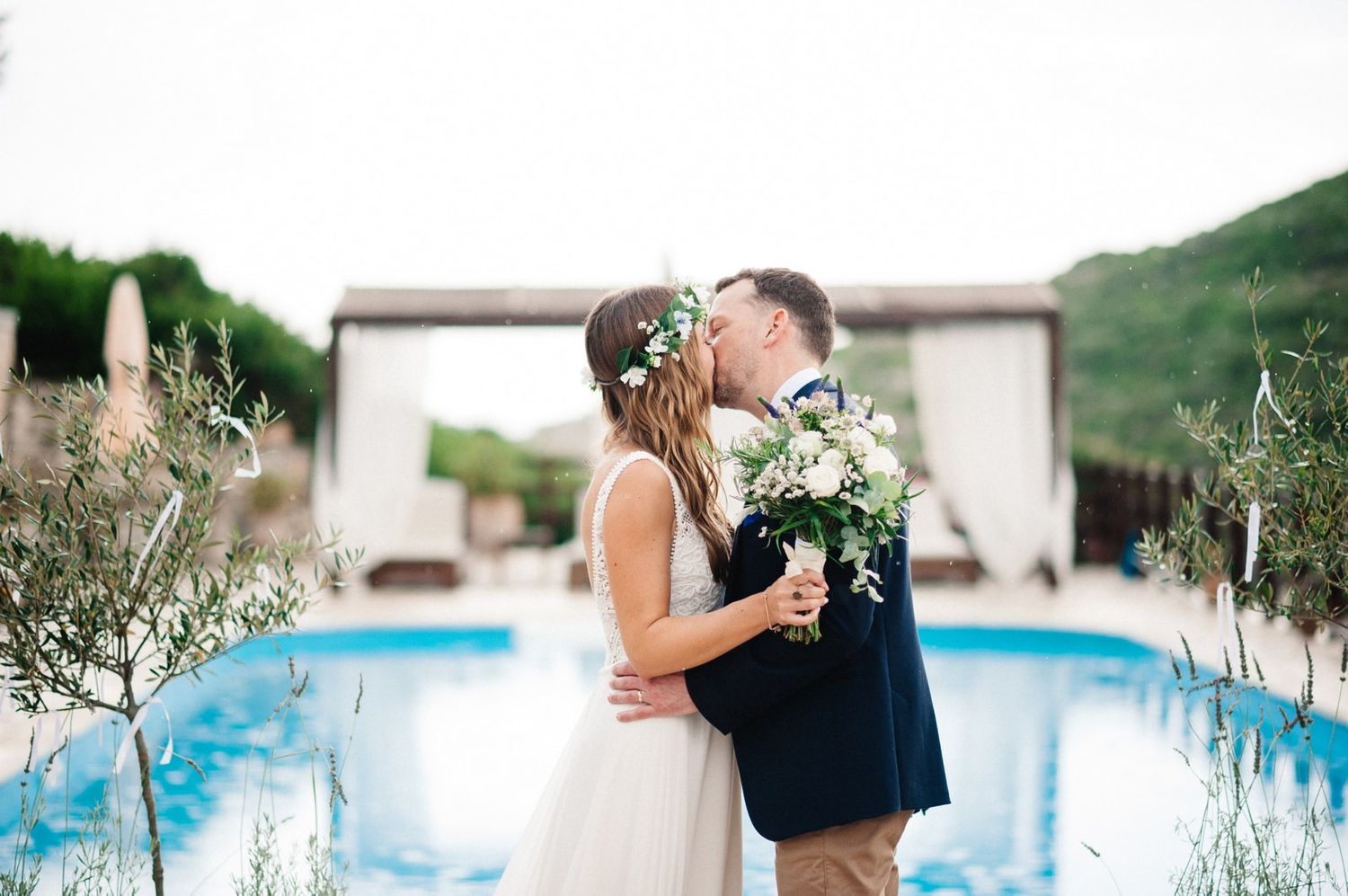 Romantic wedding kiss by pool with stone archway and mountain backdrop.