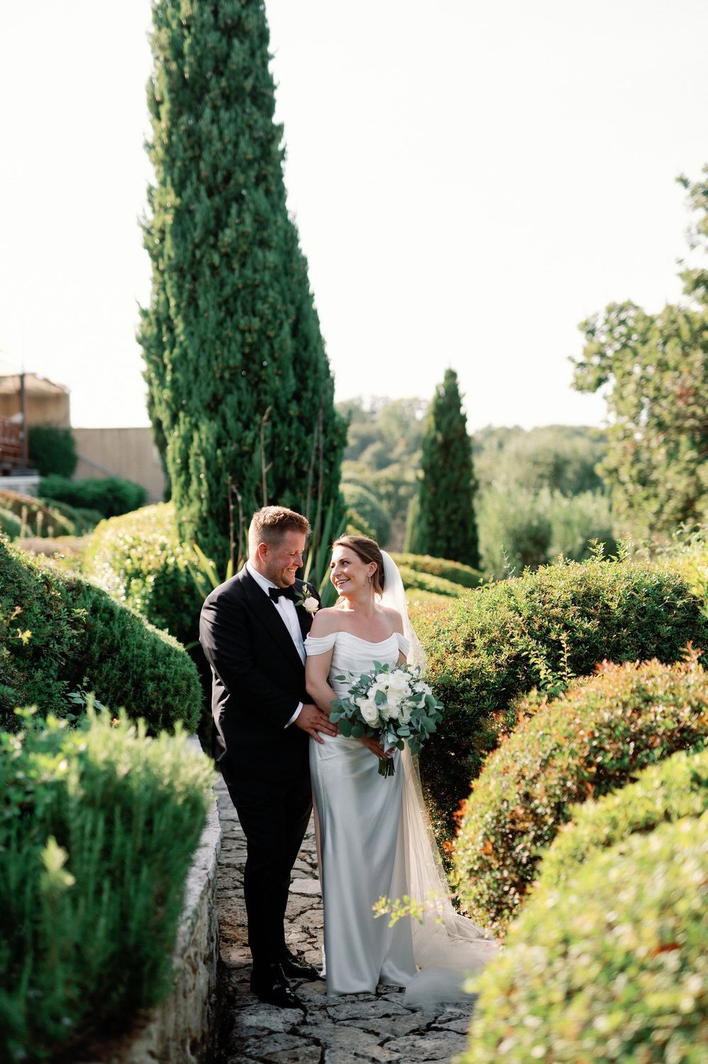 A wedding couple stands on a stone path surrounded by lush cypress trees and greenery in at Cavo Alkyoni in Corfu