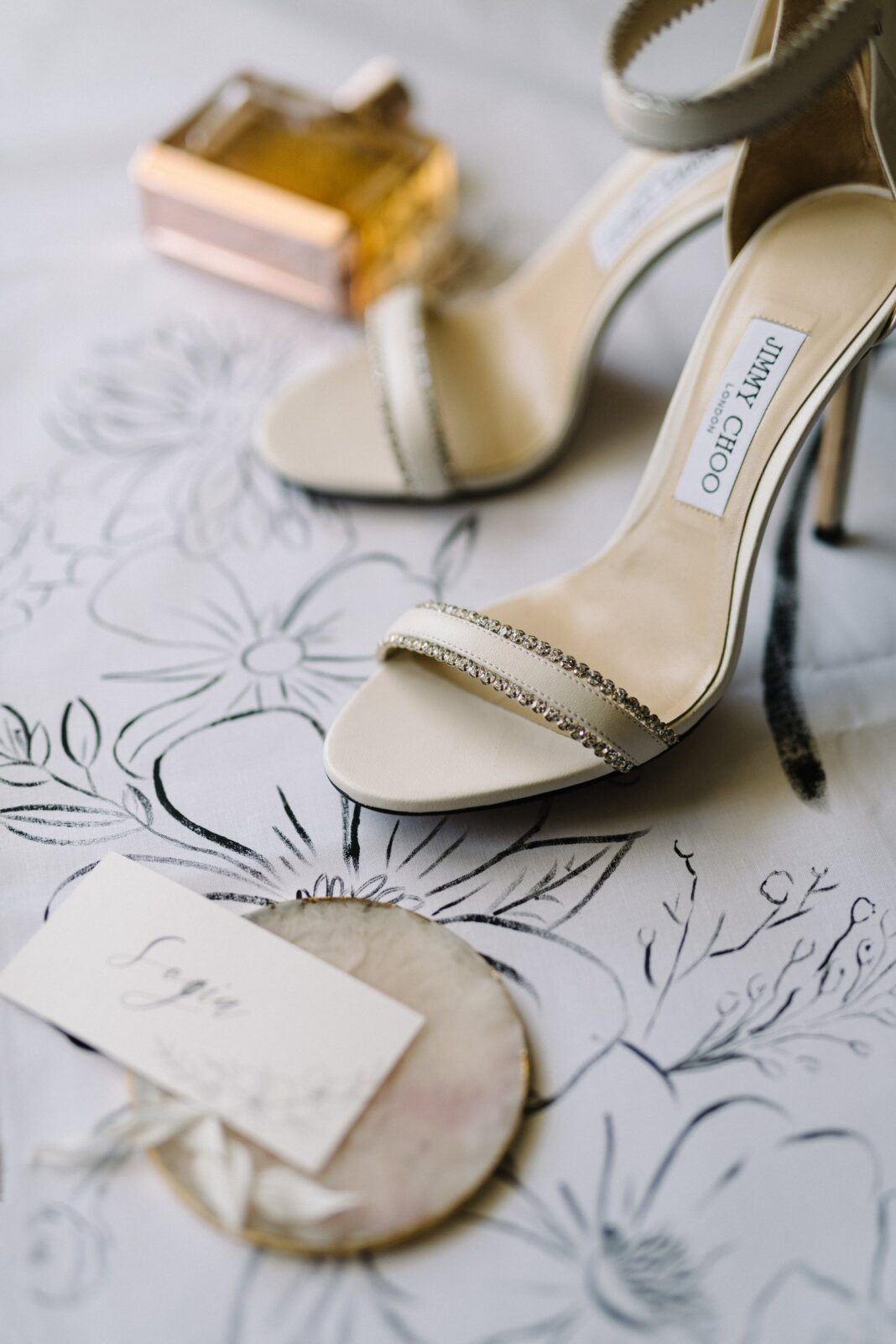 Elegant Jimmy Choo bridal sandals with crystal straps displayed on floral patterned paper beside a perfume bottle.