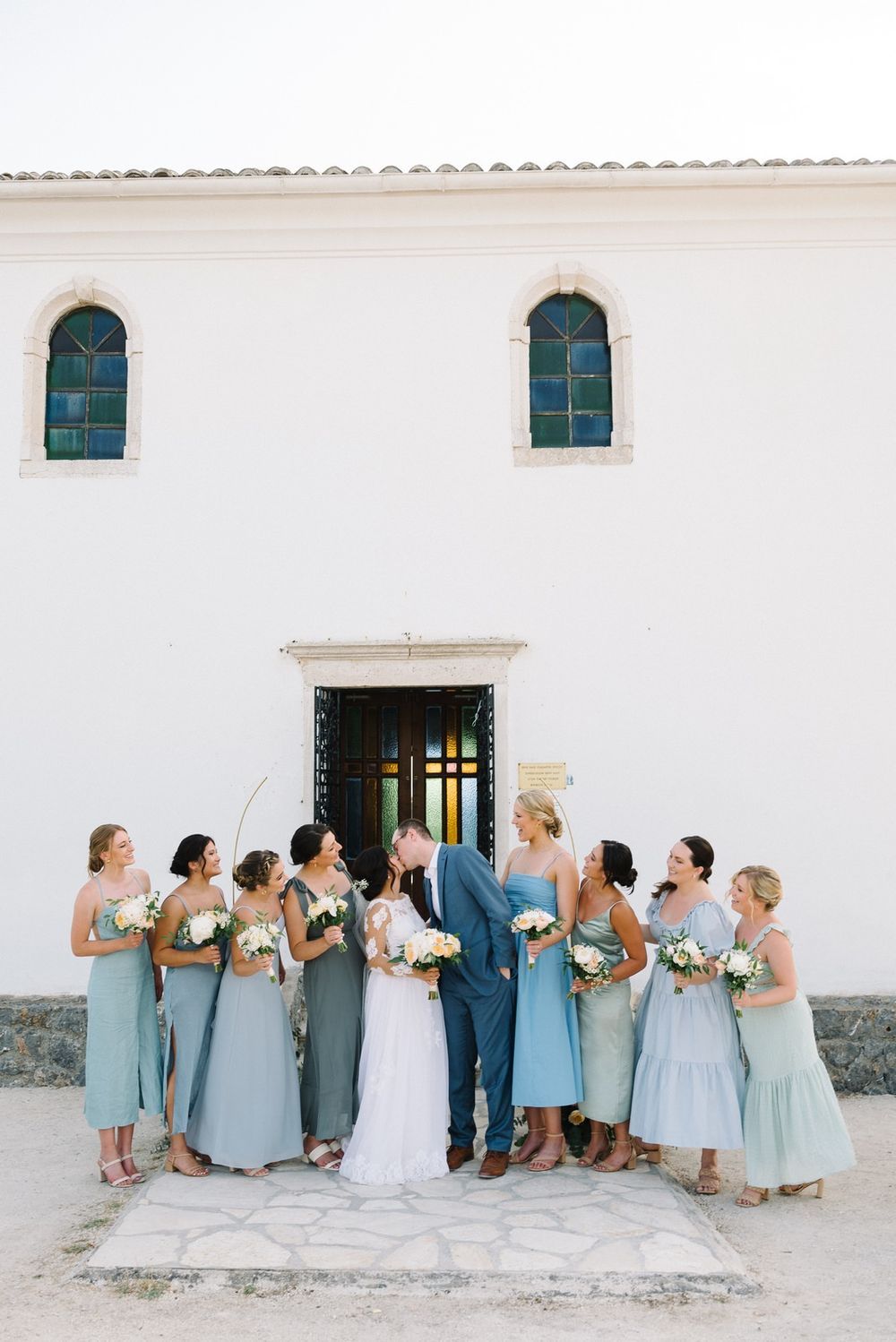 Wedding party in light blue bridesmaid dresses lined up against a white building wall for group photos.