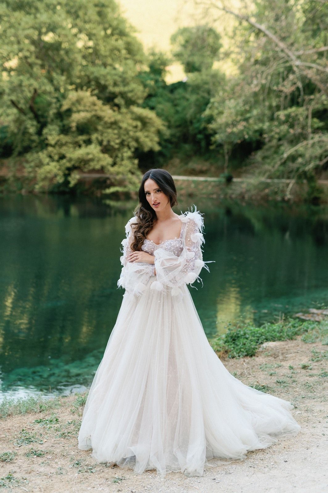 Ethereal white wedding gown with feathered sleeves and flowing tulle skirt photographed against tranquil turquoise lake.