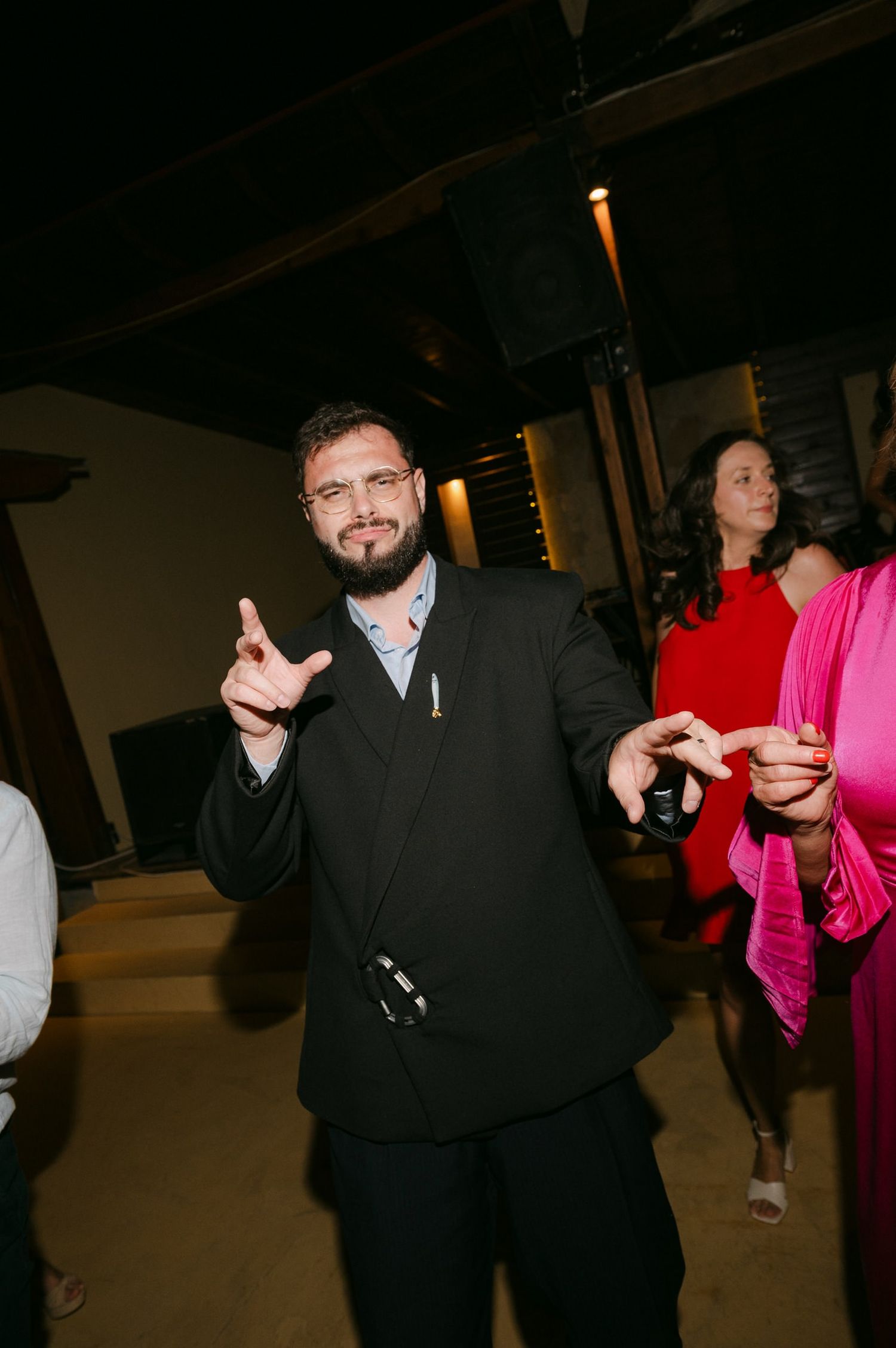 A person in a black suit gestures at a social gathering while others mingle in the background.