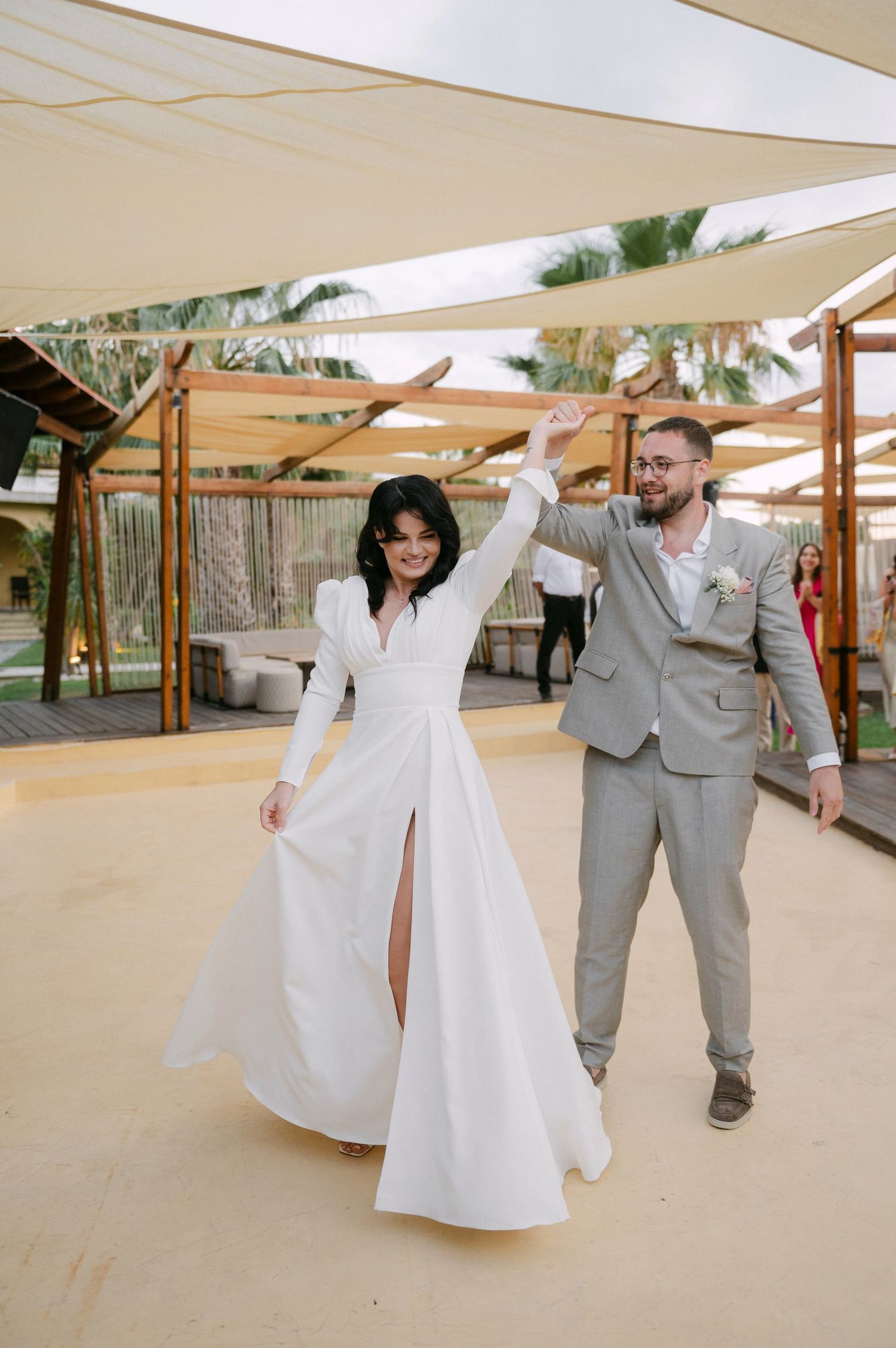 A couple in wedding attire dances and twirls together on an outdoor patio with palm trees in the background.