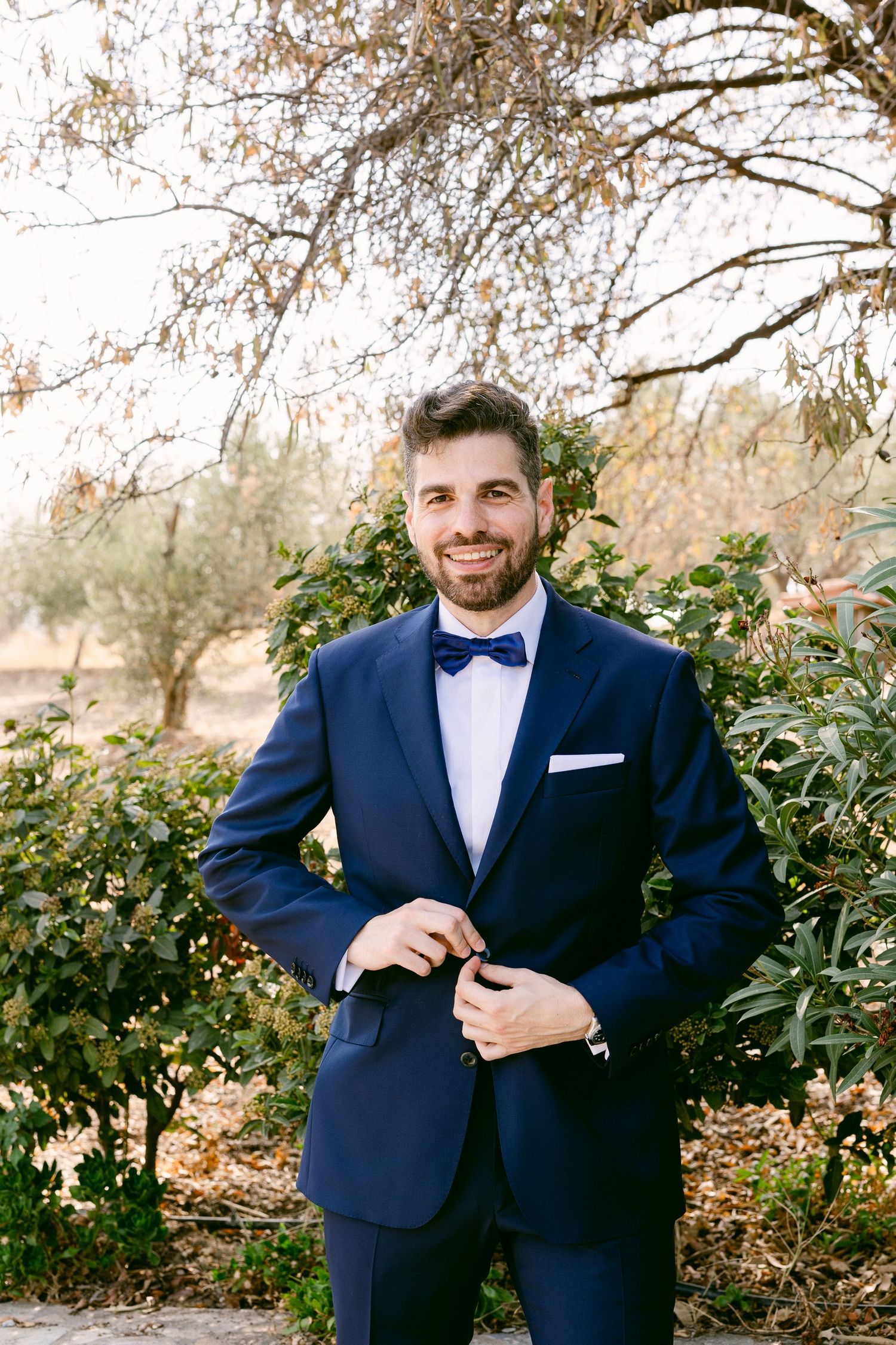 A person in a navy blue tuxedo with bow tie poses outdoors among autumn foliage in a formal portrait setting.
