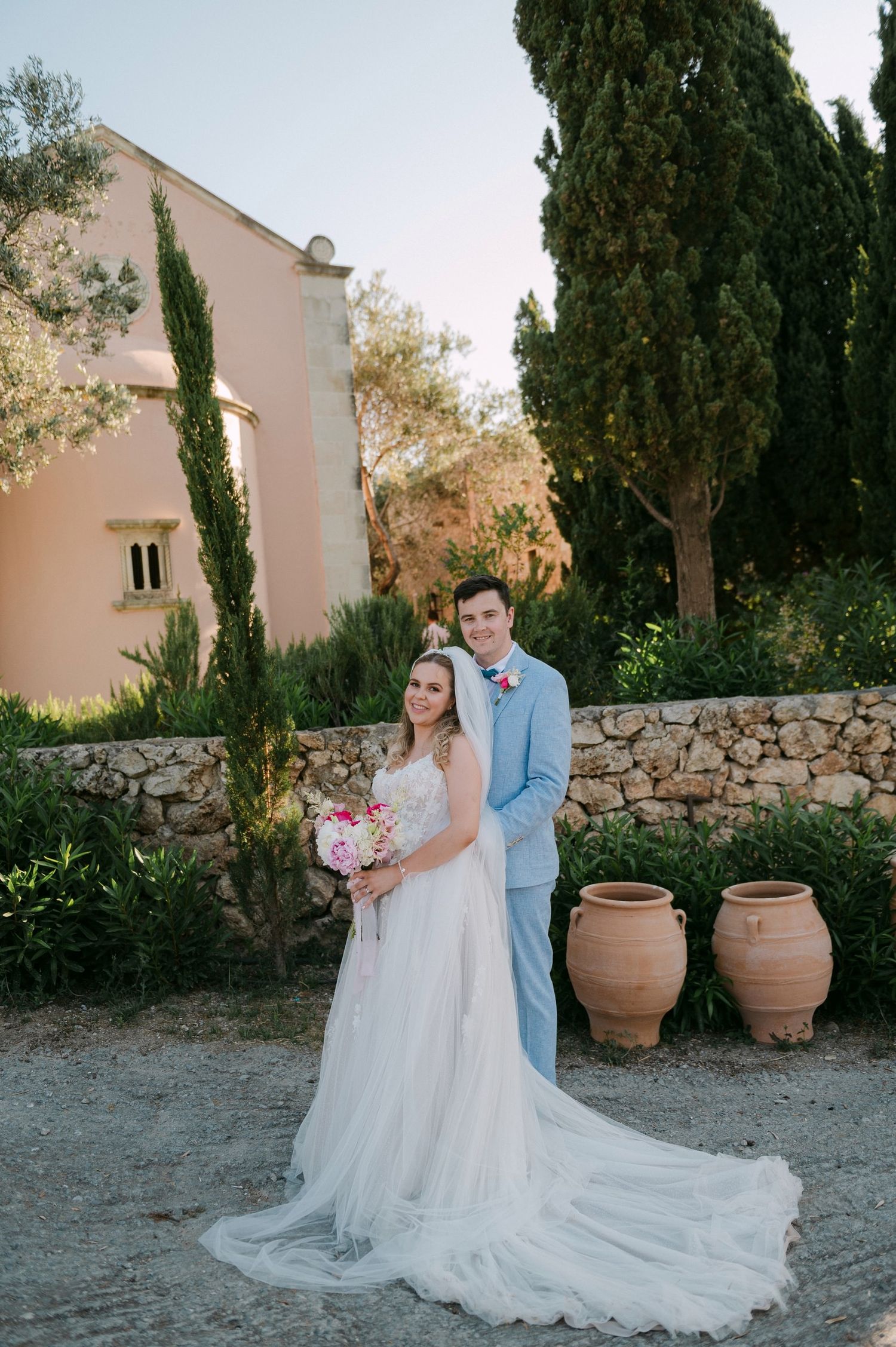 Wedding couple poses in Mediterranean garden setting with cypress trees and stone wall for romantic outdoor portraits.