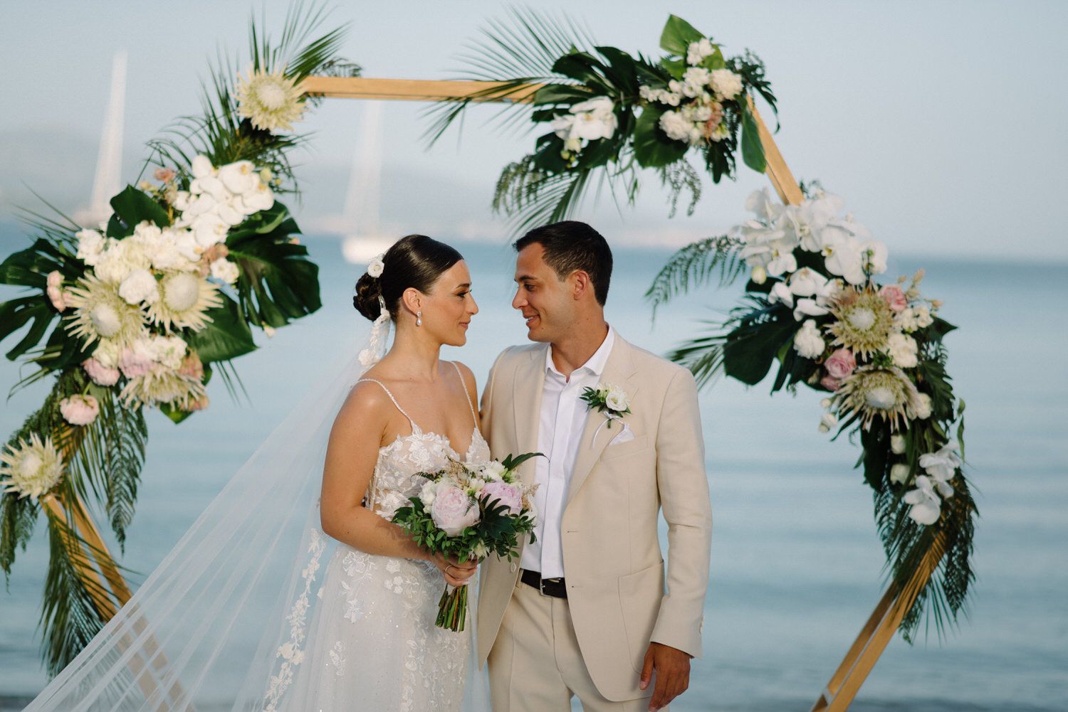 Couple shares intimate moment under floral wedding arch with ocean backdrop.