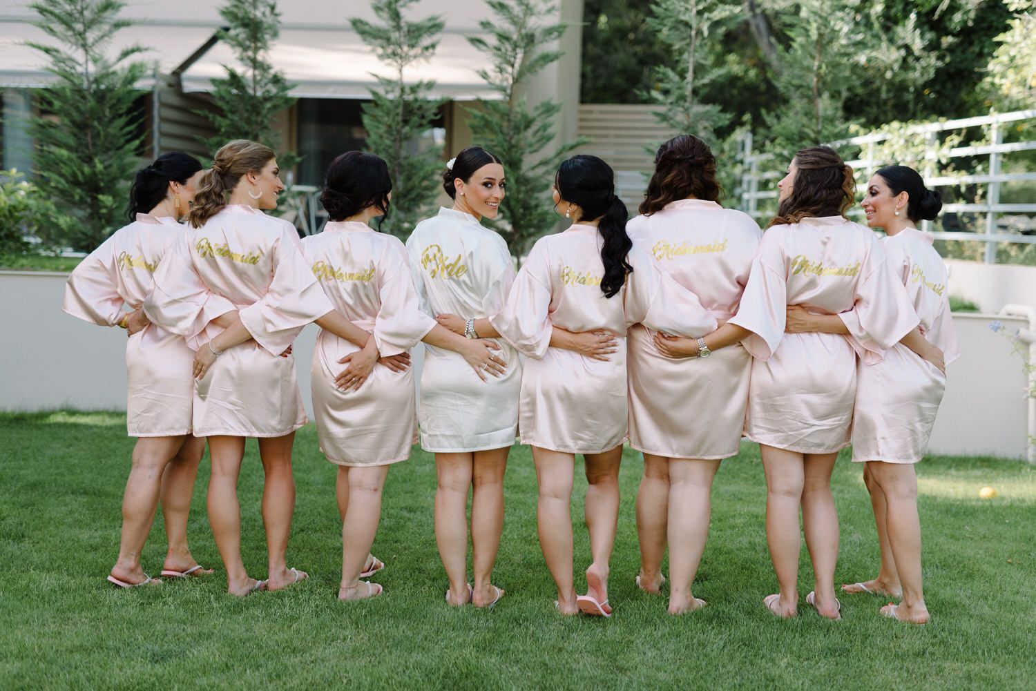 Bridesmaids stand with arms around each other showing backs of matching pink robes.