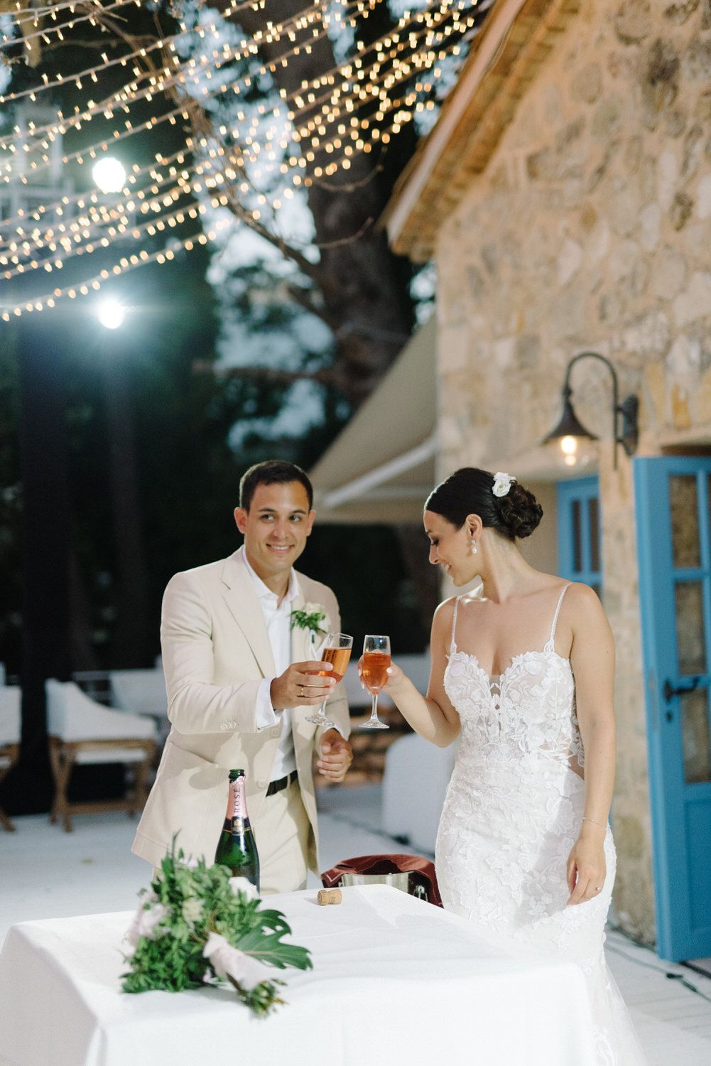 Couple toasts with champagne glasses at their wedding reception by a rustic stone wall.