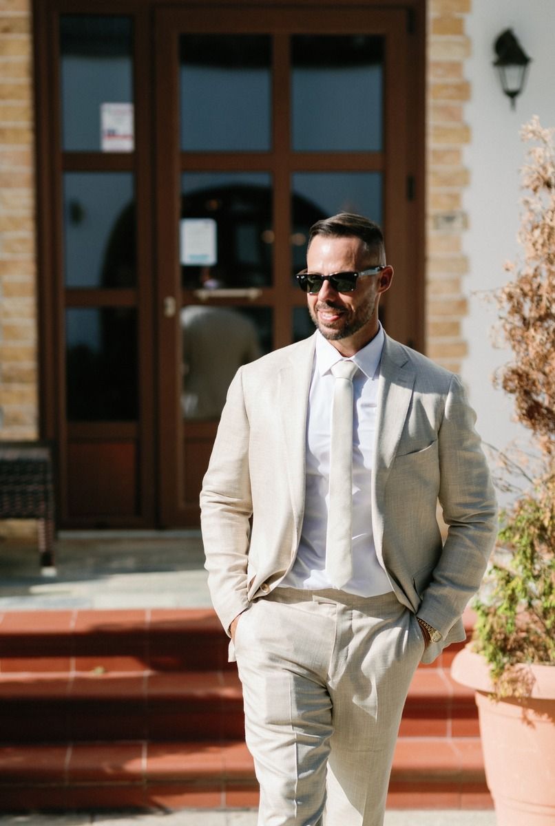 Elegant groom in light beige suit stands on brick steps with sunglasses.