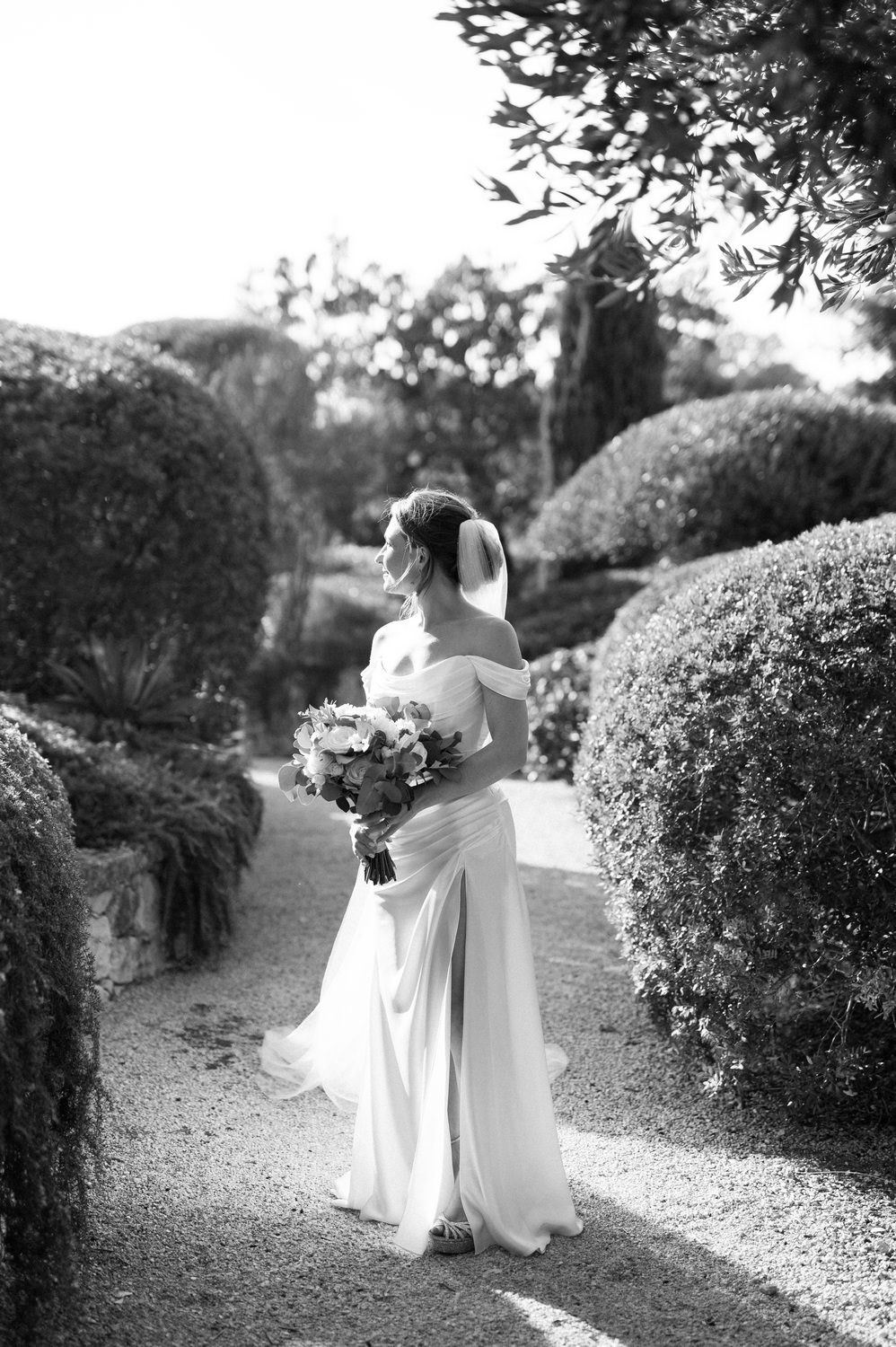 Black and white portrait of bride with bouquet on garden path surrounded by manicured hedges.