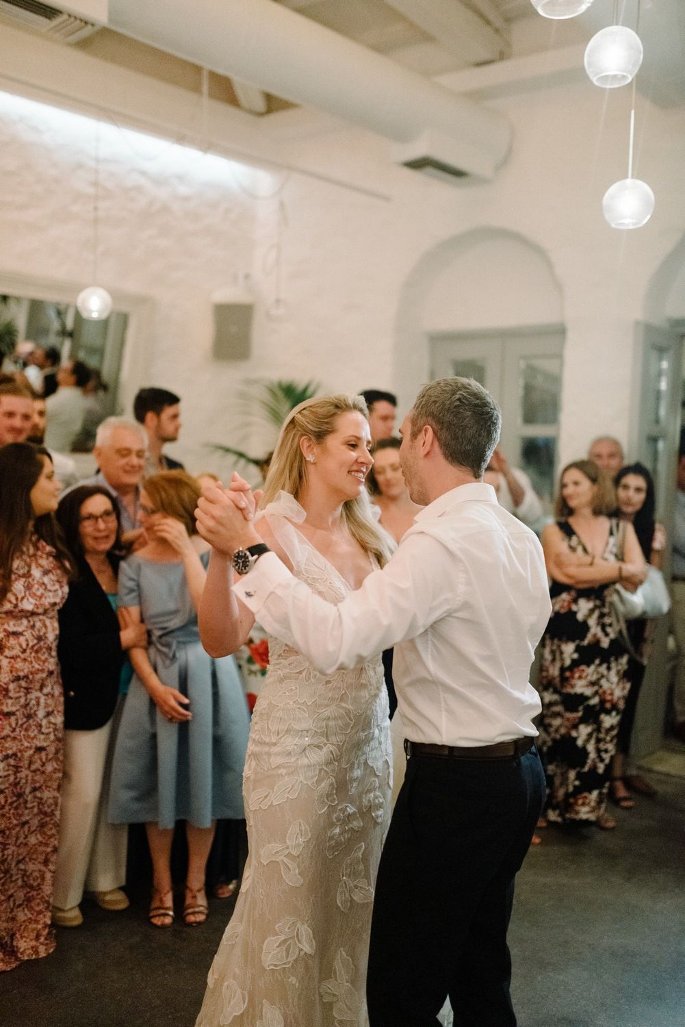 Newlyweds share their first dance surrounded by applauding guests in an elegant white-walled venue.