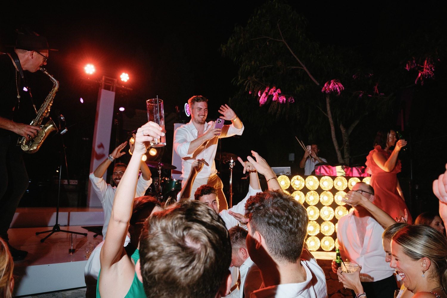 Crowded dance party scene with performers and audience against backdrop of yellow lights.