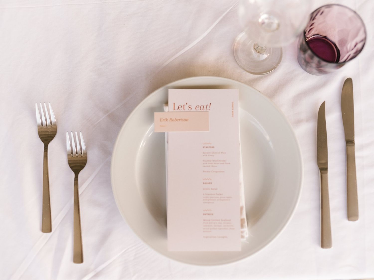 Elegant place setting with gold cutlery, menu card and wine glass on white tablecloth.