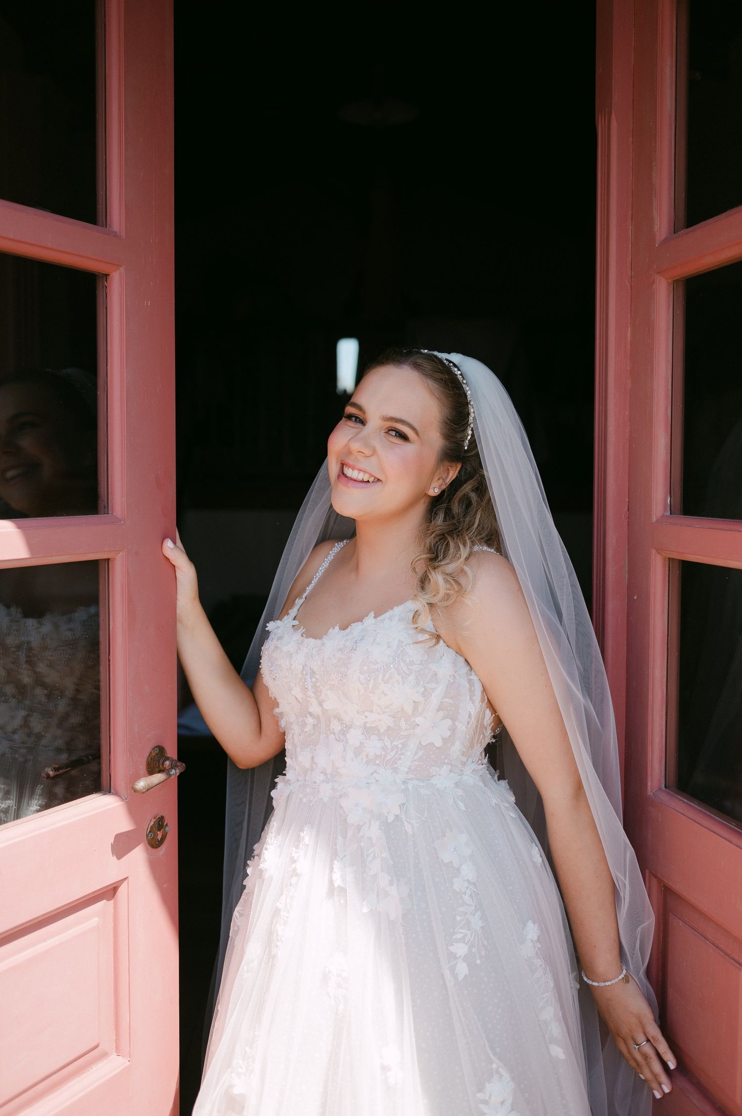 A bride in a white lace wedding dress laughs joyfully while standing in front of pink-framed windows.